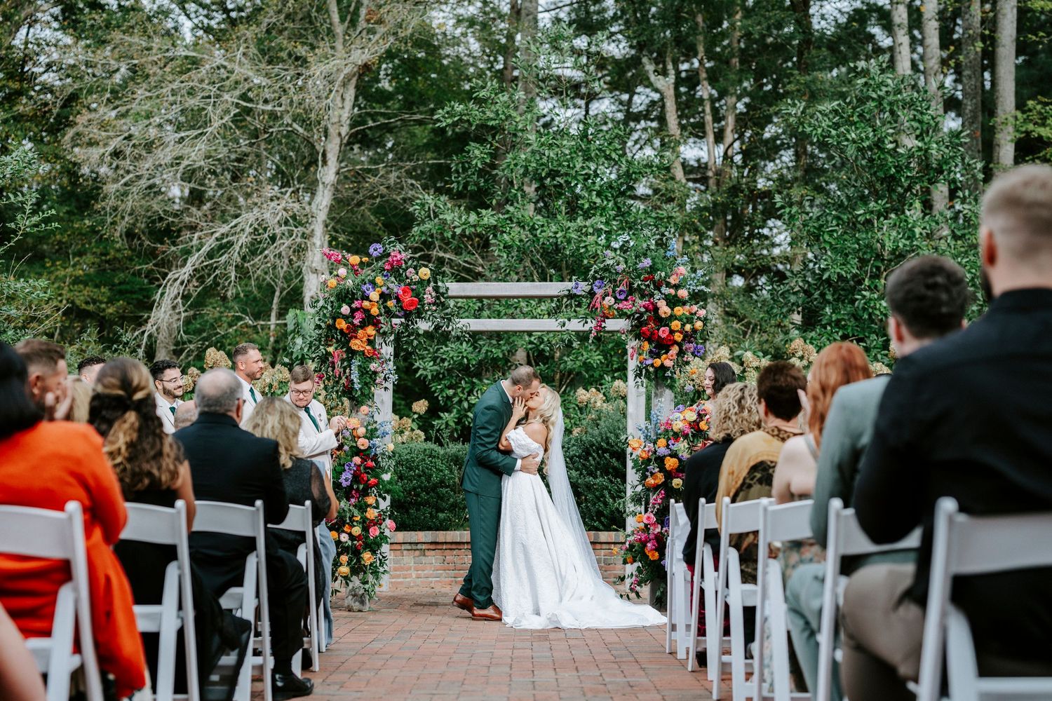An outdoor garden wedding ceremony under a floral arch with guests seated on white chairs along a brick pathway.