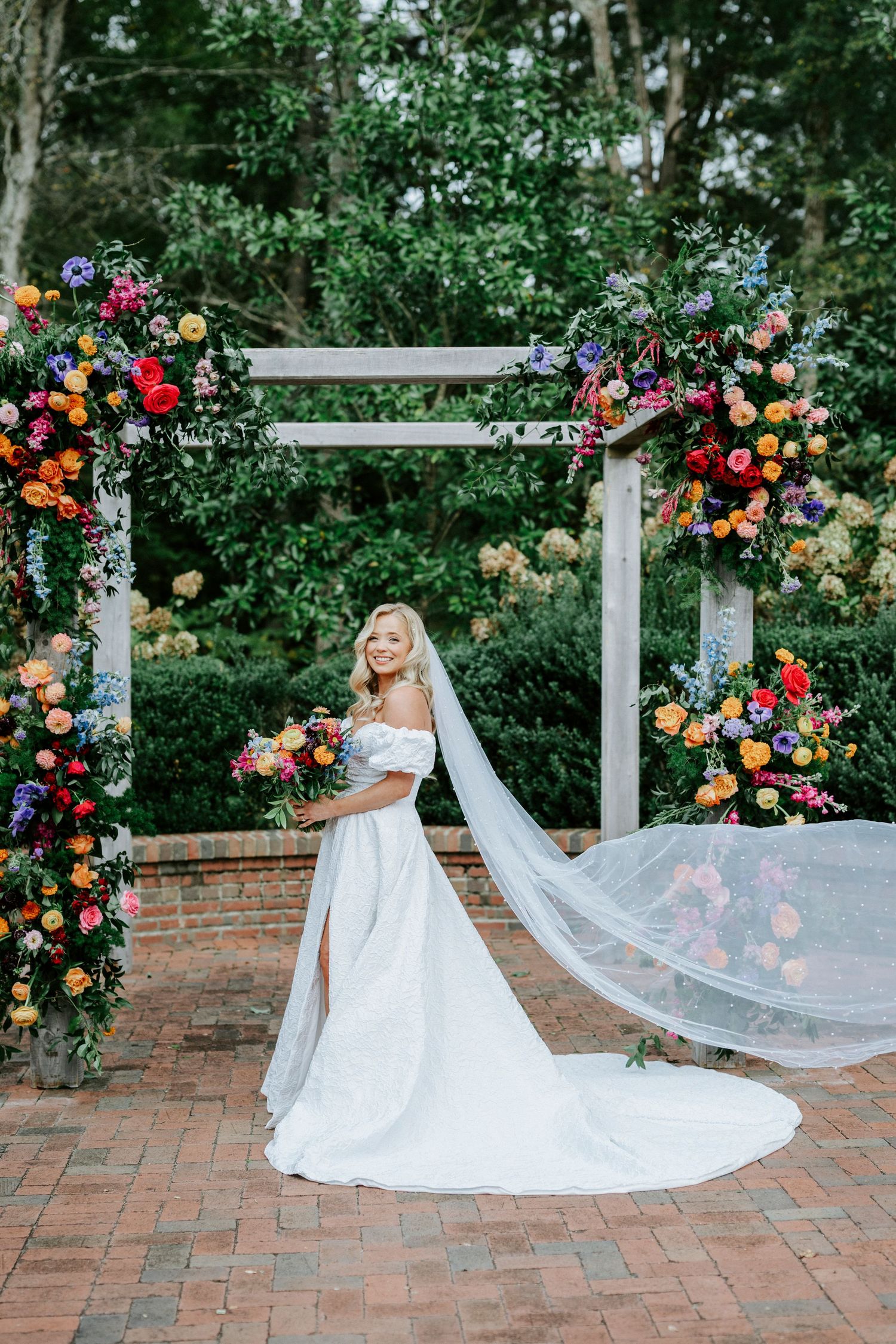 A flowing white wedding dress and veil cascade across brick pavers beneath a floral-adorned garden arch.