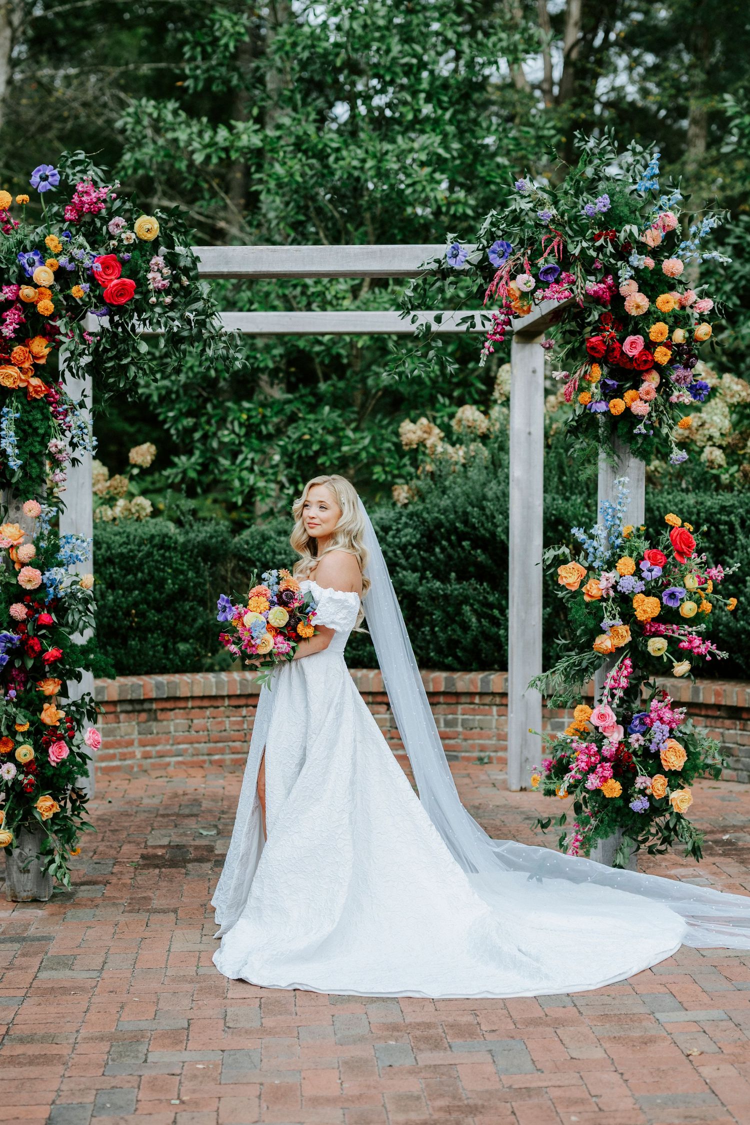 Colorful floral arrangements adorn a white garden arch over a brick patio with a sweeping bridal gown.