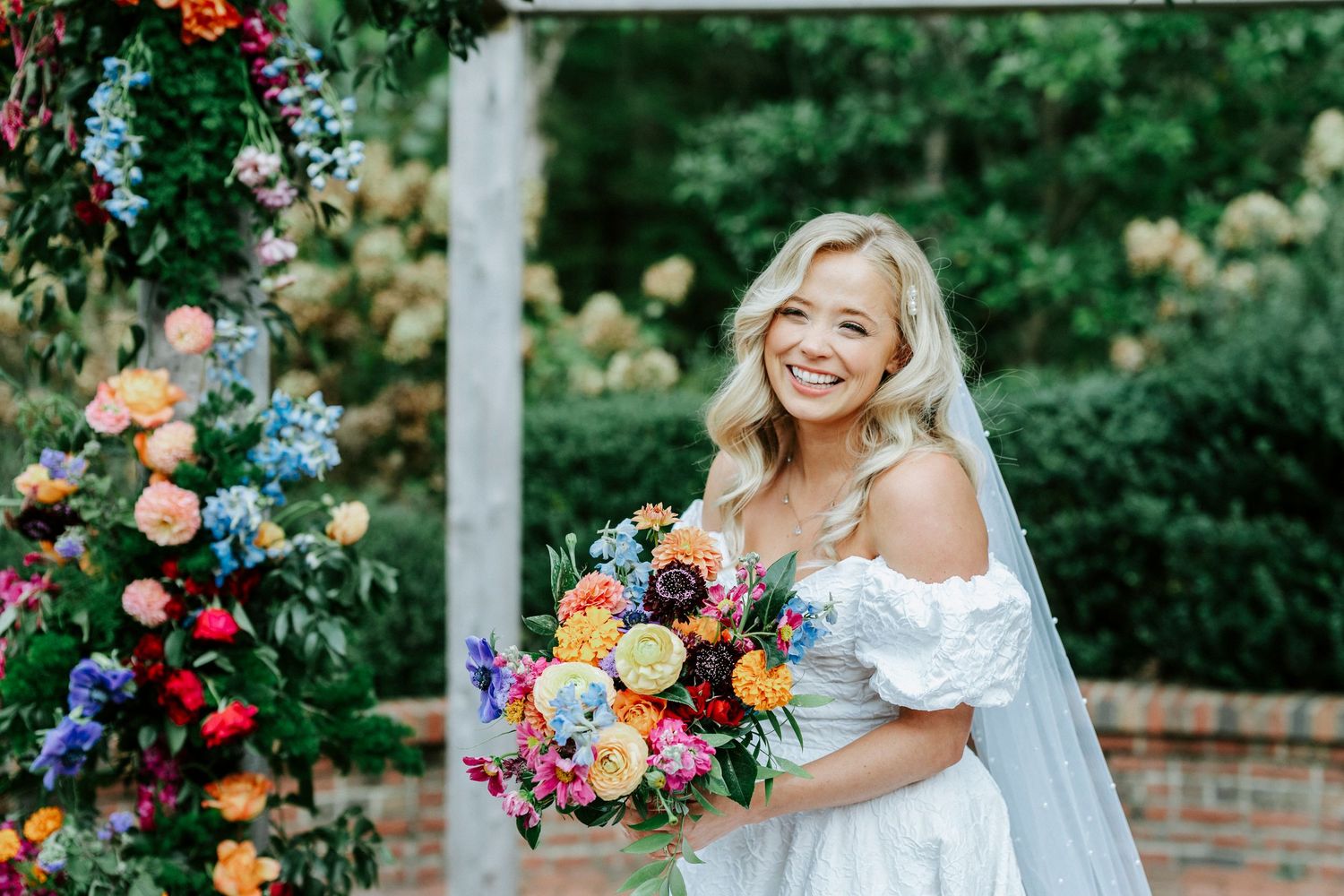 A bride in a white dress poses with a vibrant colorful bouquet of flowers in an outdoor garden setting.