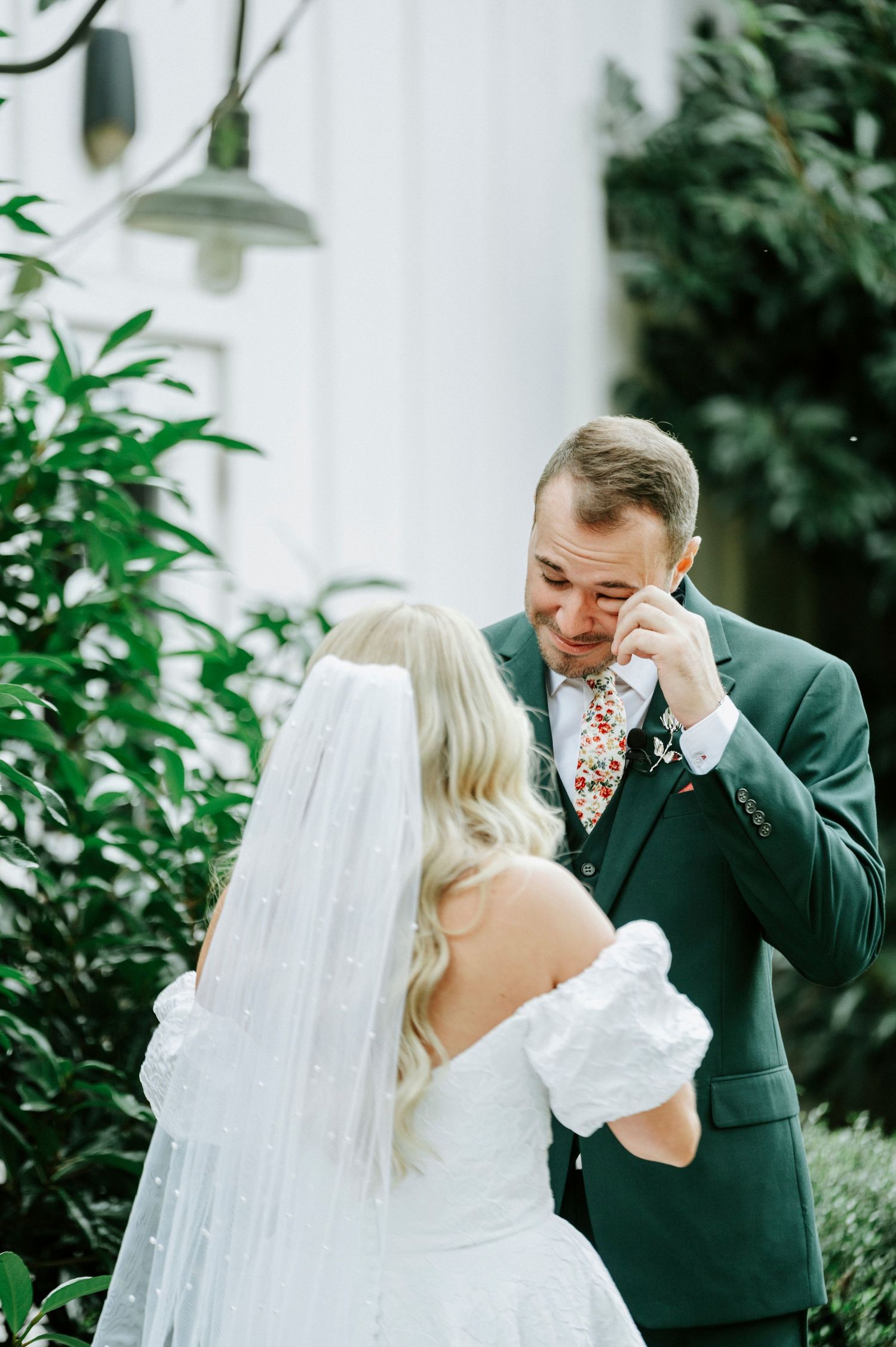 A couple in wedding attire share an emotional moment in a garden setting with white veil and green foliage.