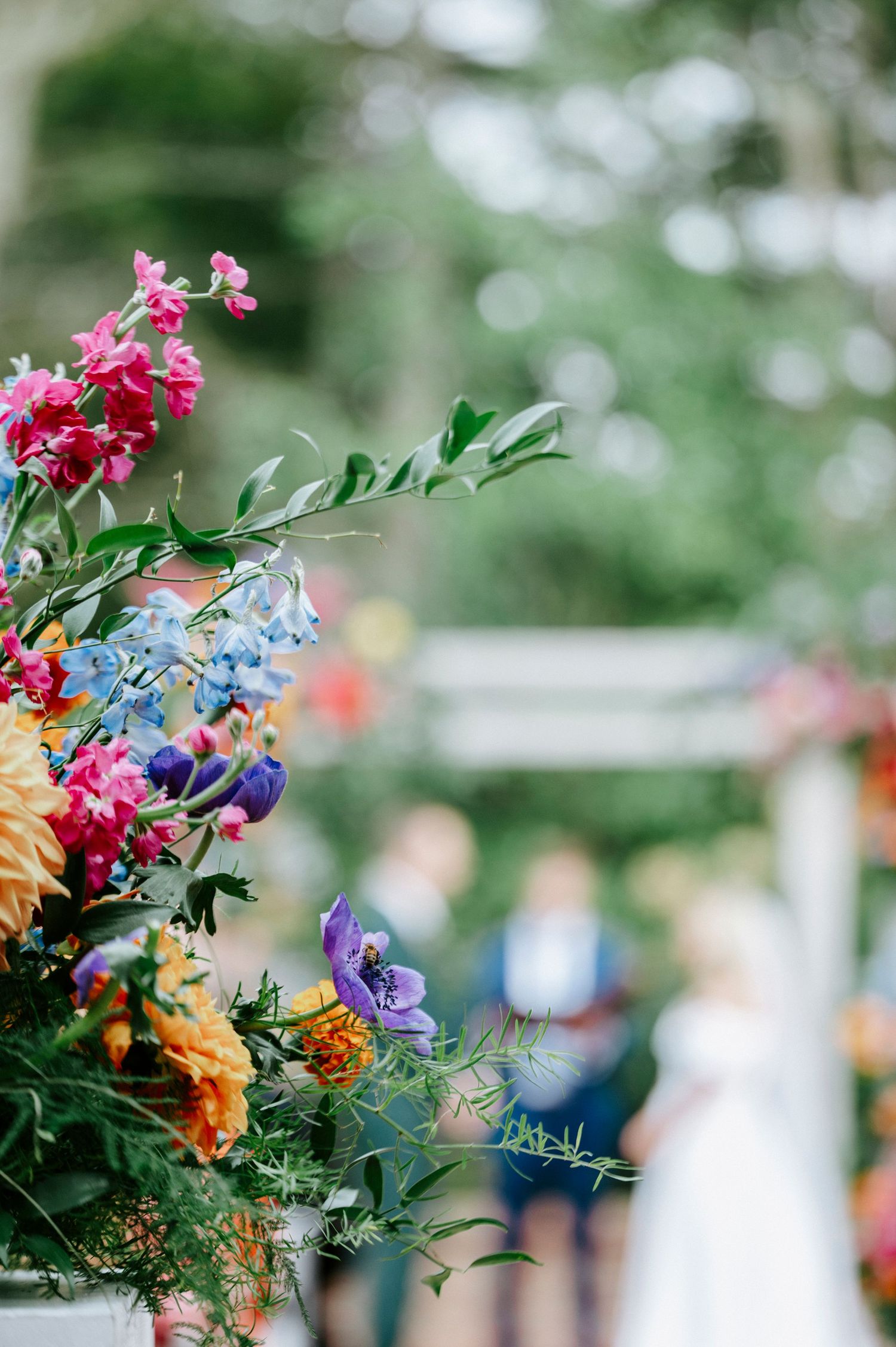 Colorful garden flowers including pink, blue, and yellow blooms arranged in a decorative outdoor display with soft bokeh background.