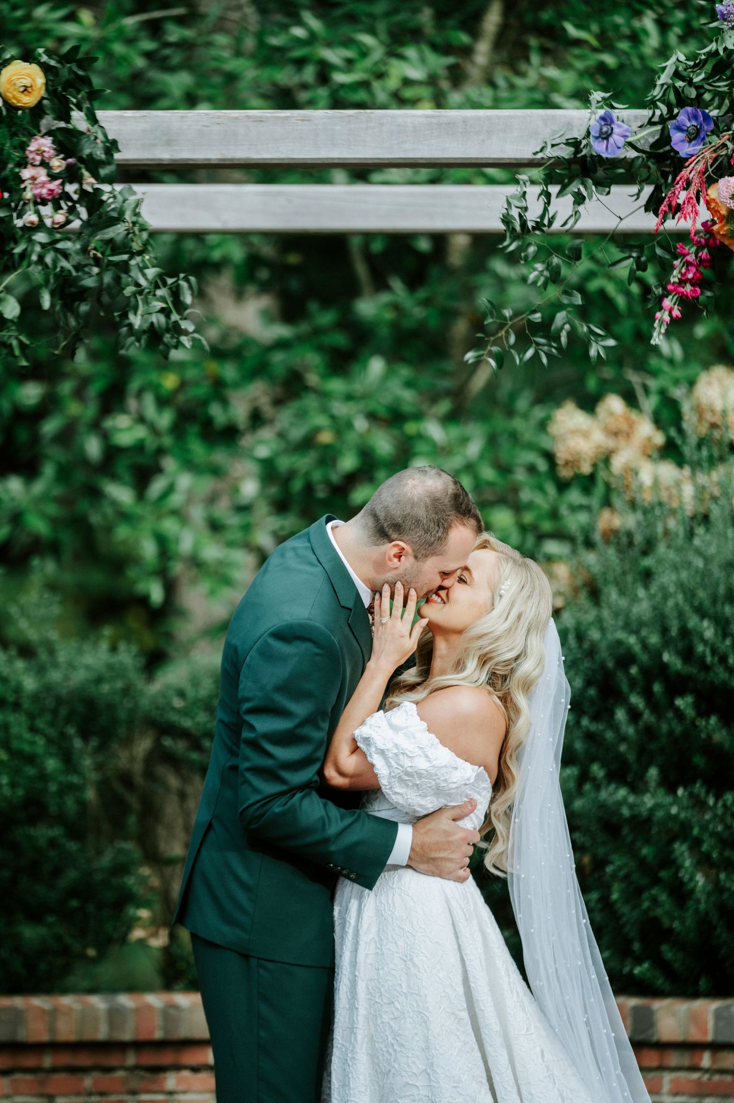 A bride and groom share romantic kisses in a lush garden setting with brick walls and colorful flowers.