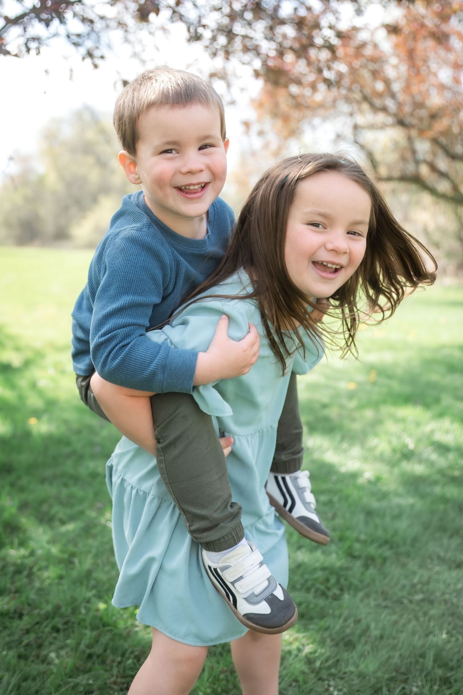 Two children playing and laughing together outdoors in a piggyback pose during autumn with fall foliage in background.