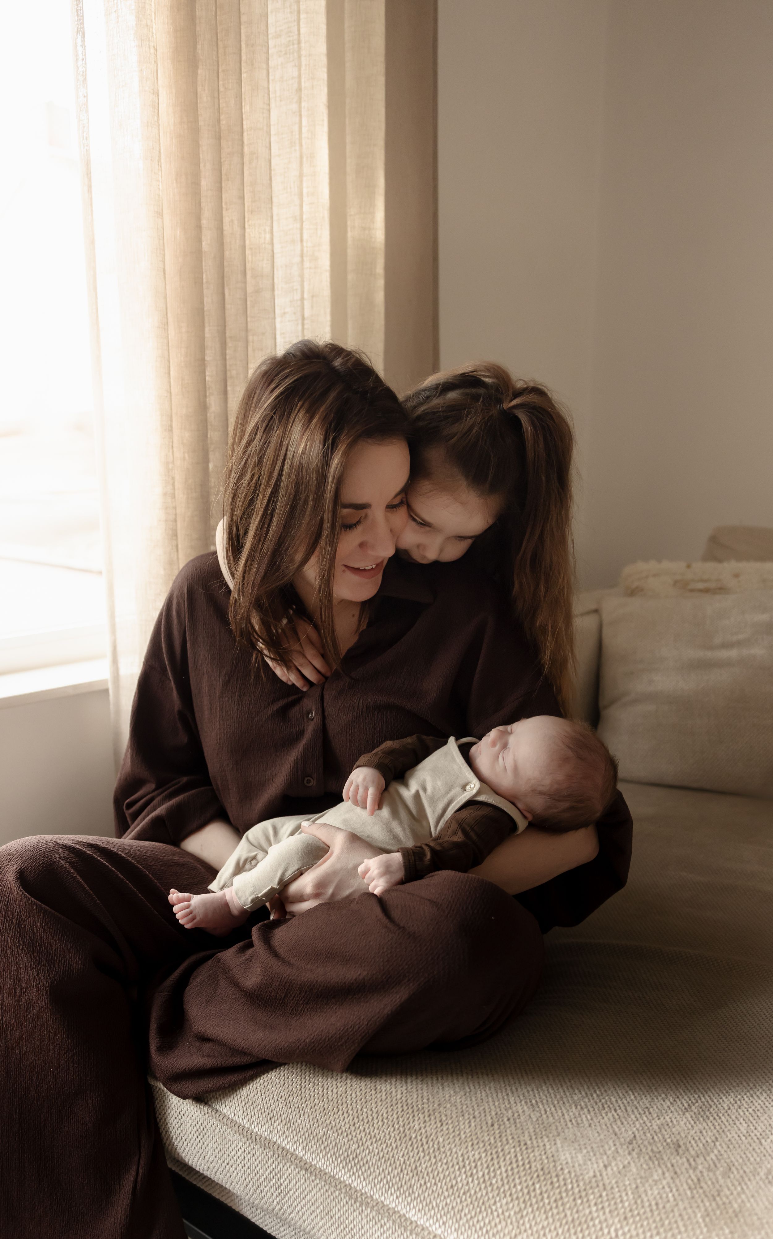 A tender moment between caregivers and newborn baby on a couch in warm brown tones.