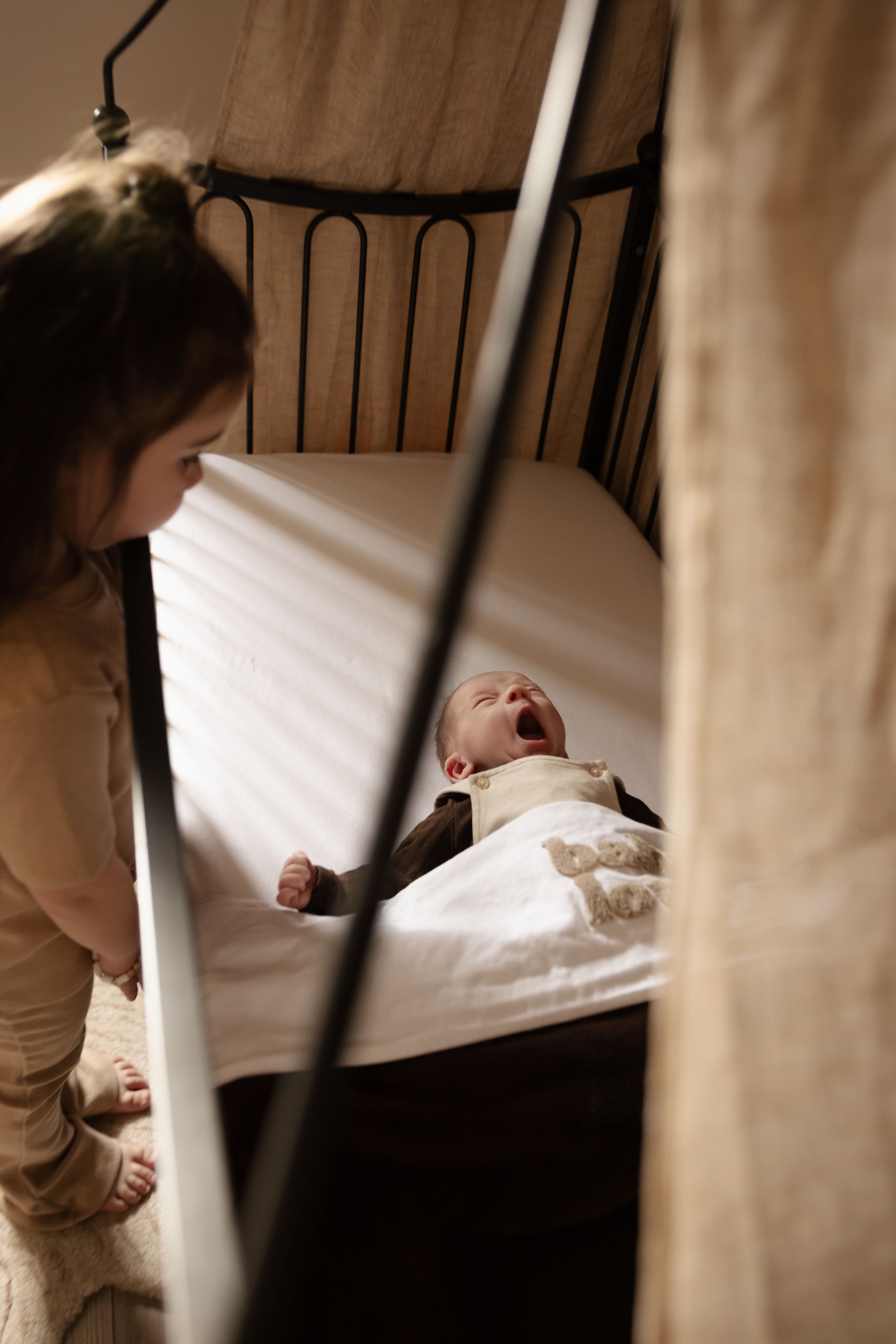 A baby lying in a crib laughs while being watched through the bars.