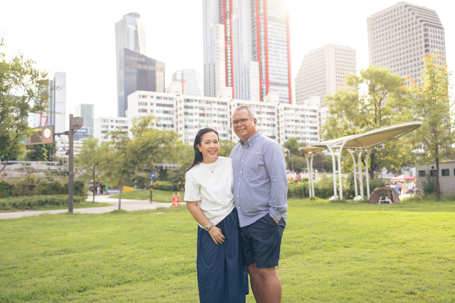 Happy couple walking together and laughing in an urban park with tall buildings in the background on a sunny day.