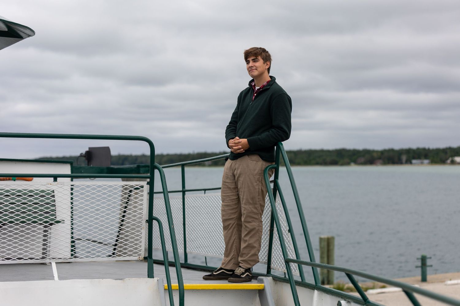 Young man standing on a ferry boat deck with a cloudy sky and waterfront buildings in the background.