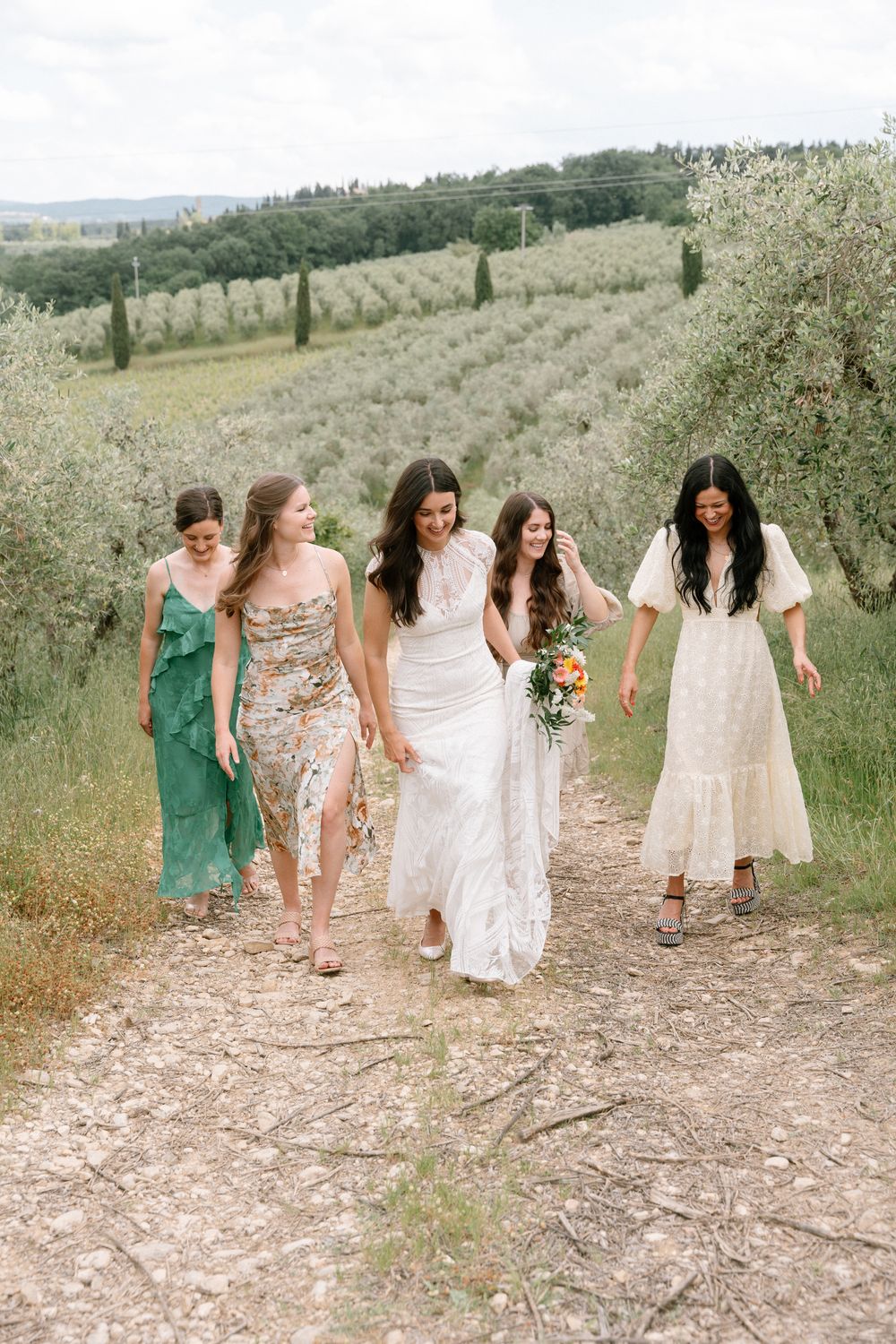 A group of women in flowing dresses walking along a rustic path through an Italian countryside.