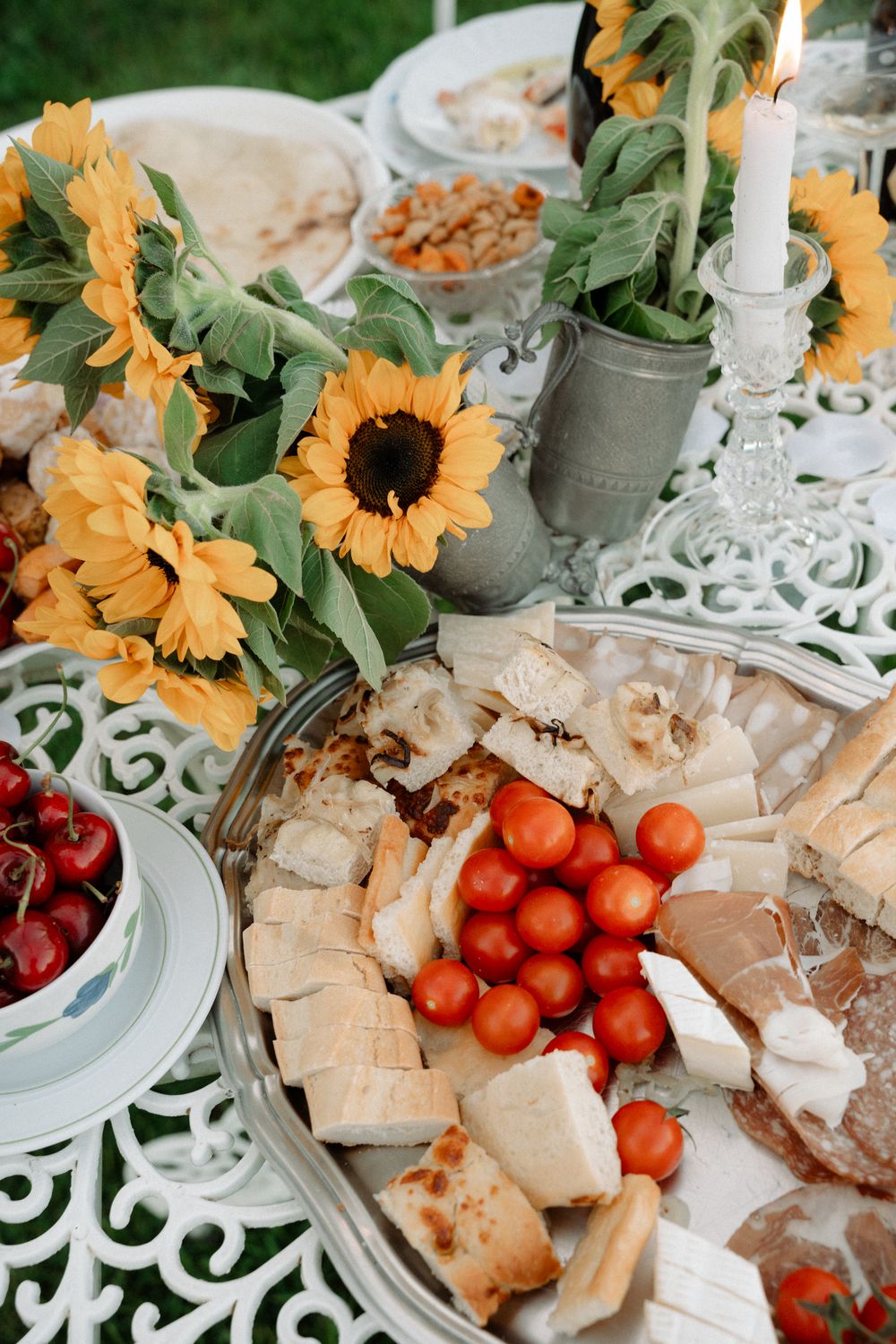A beautifully arranged party spread with sunflowers, tomatoes, sandwiches and cherries on an ornate white table.