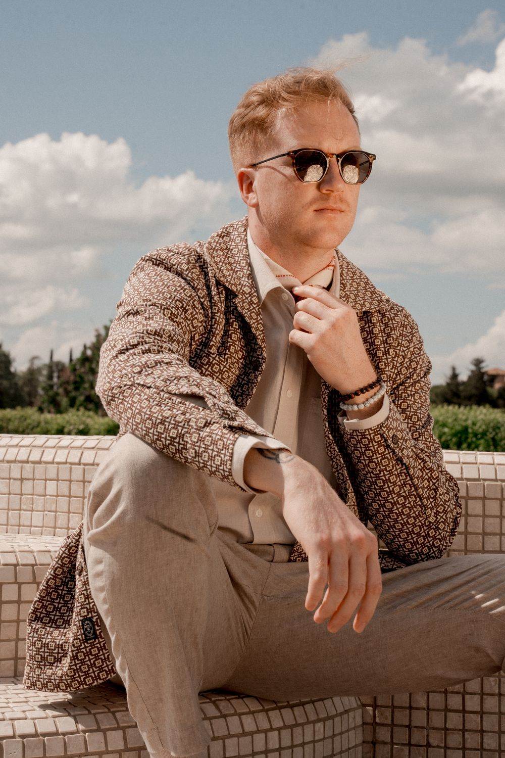 Person in brown sweater and sunglasses posing thoughtfully on concrete ledge.