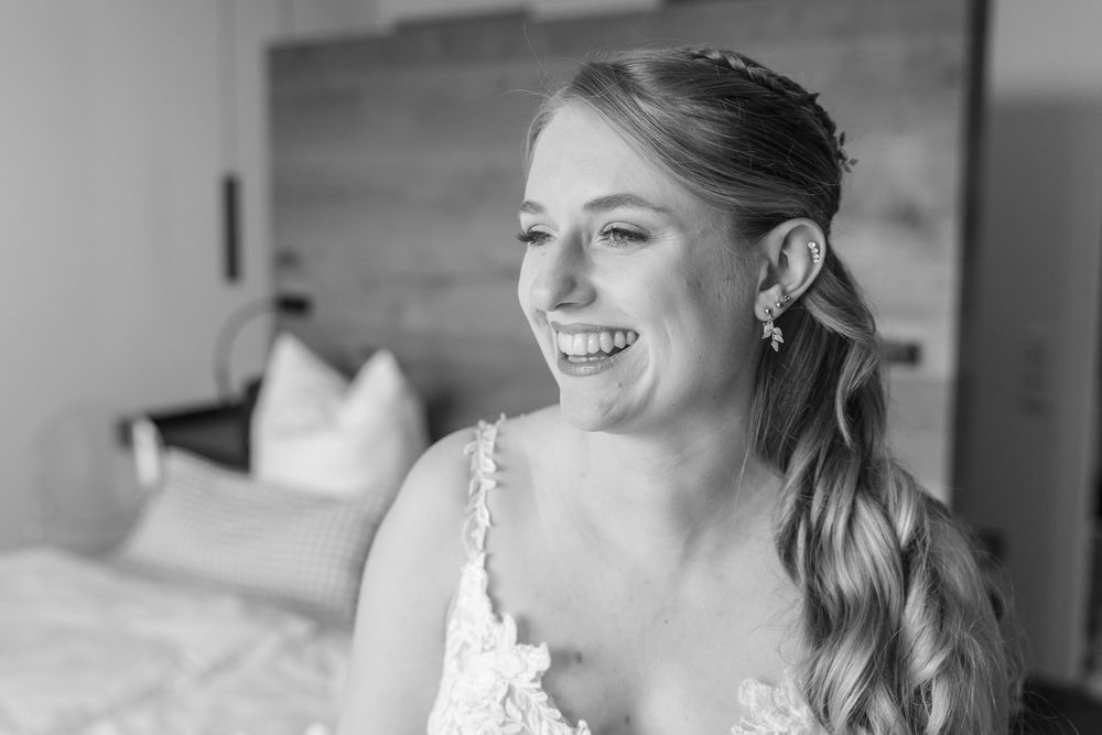 A bride in a white lace wedding dress smiles joyfully while getting ready for her wedding day.