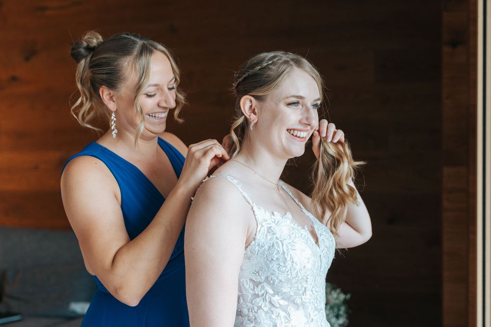 A bridesmaid in a blue dress helps adjust the white lace wedding gown during wedding preparations.