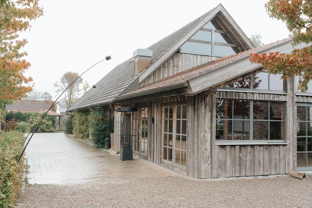 Rustic wooden barn-style building with large windows and gable roof surrounded by autumn trees on a curved pathway.