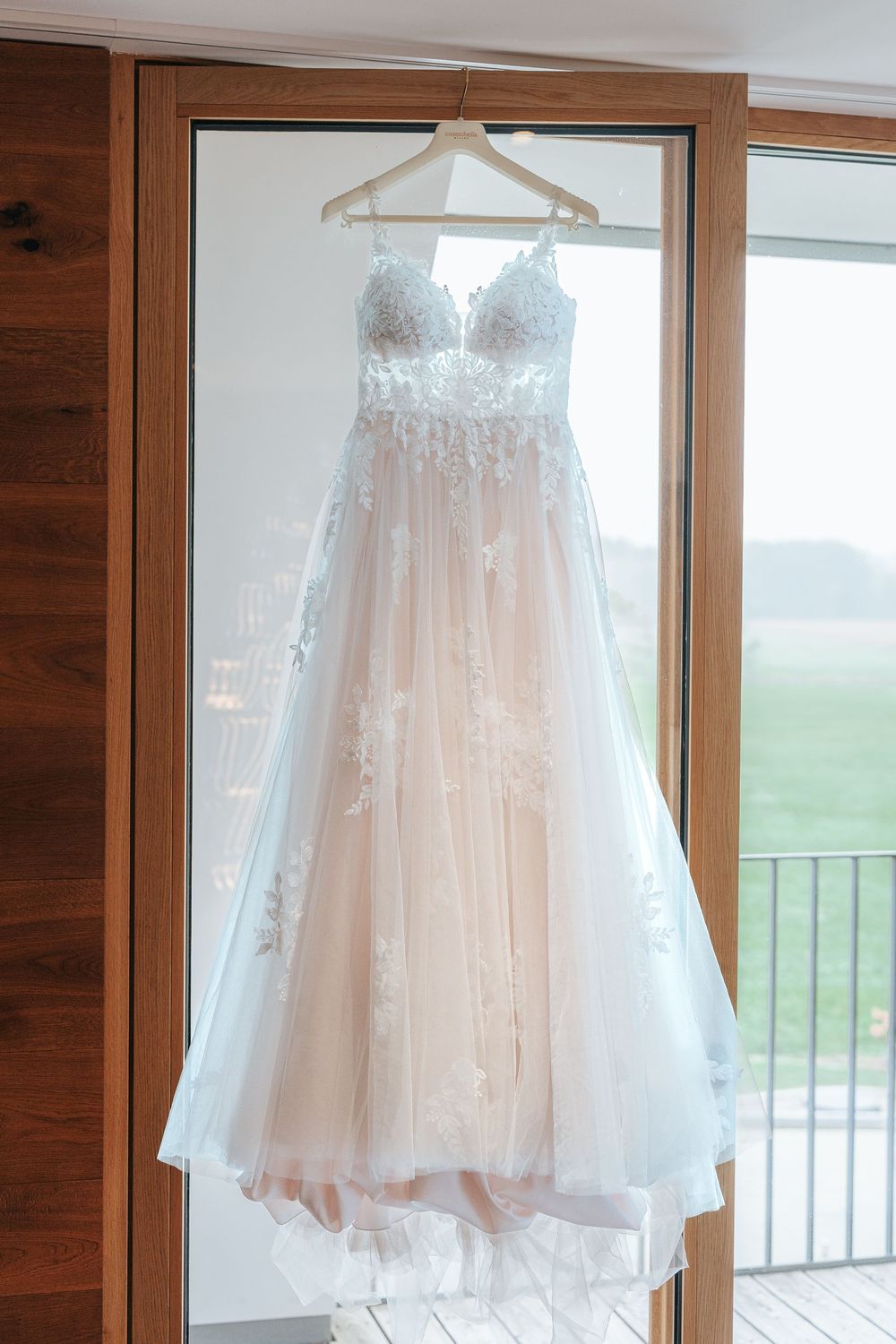 A delicate white lace wedding dress hangs from a wooden hanger by a window with natural lighting.