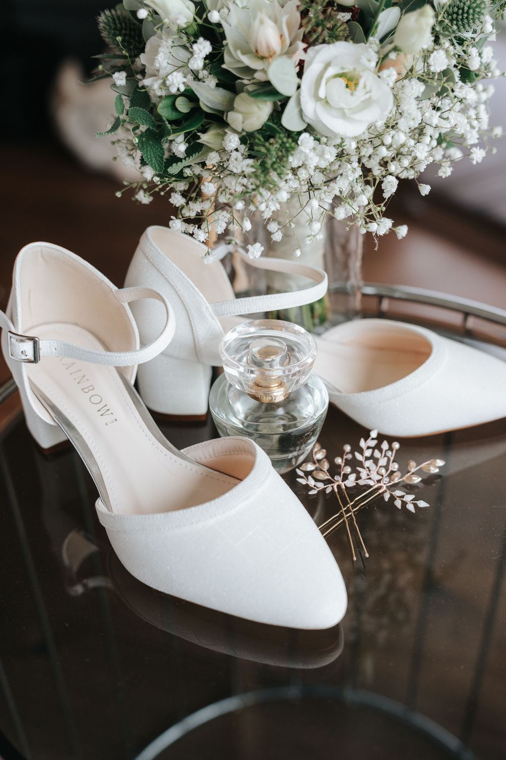 White wedding shoes with delicate floral arrangements and baby's breath in glass vases on reflective dark surface.