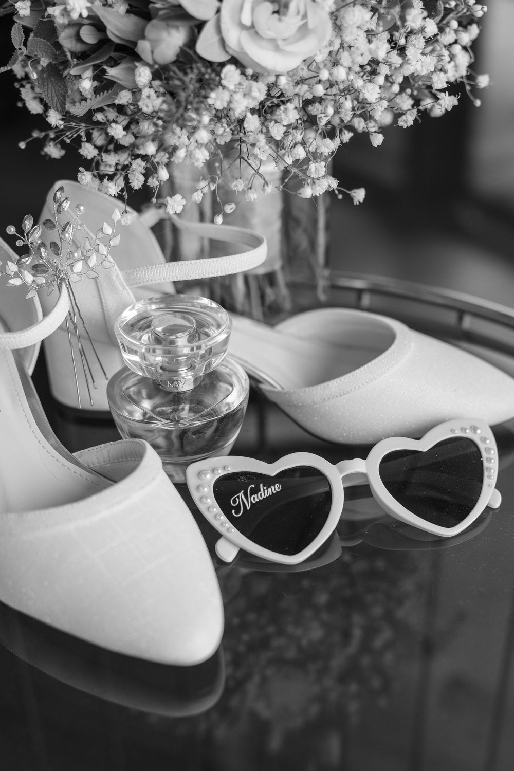 White heart-shaped sunglasses rest beside elegant bridal shoes and delicate baby's breath flowers in black and white.