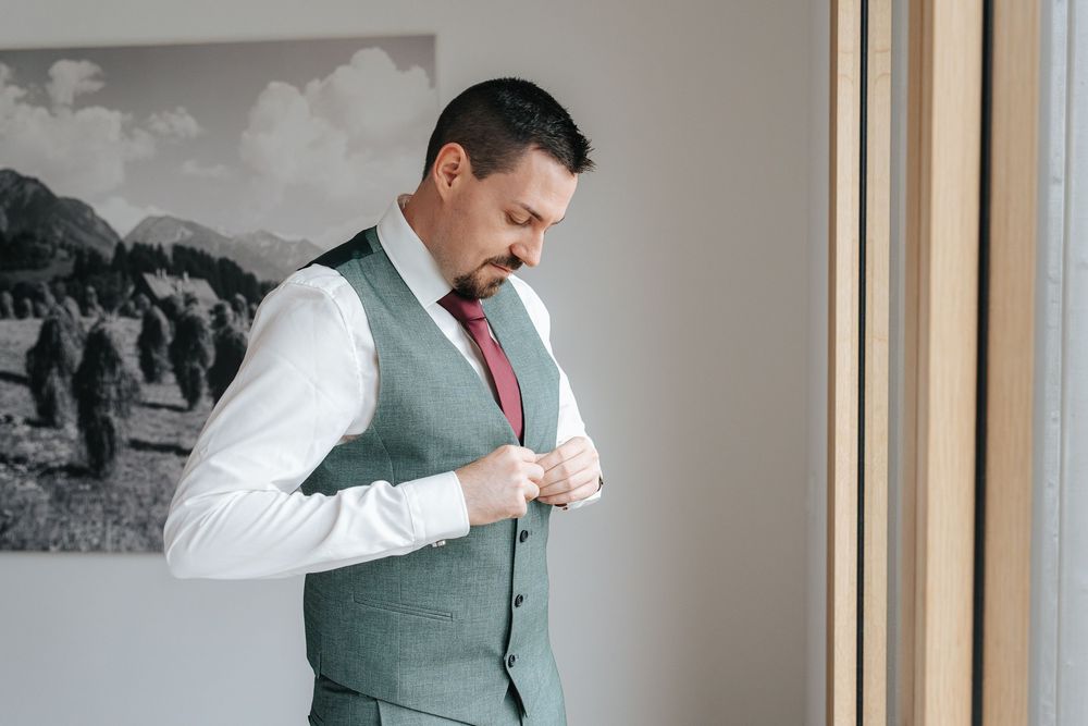 A person in a light gray vest and red tie adjusts their cuff link while standing near a window.