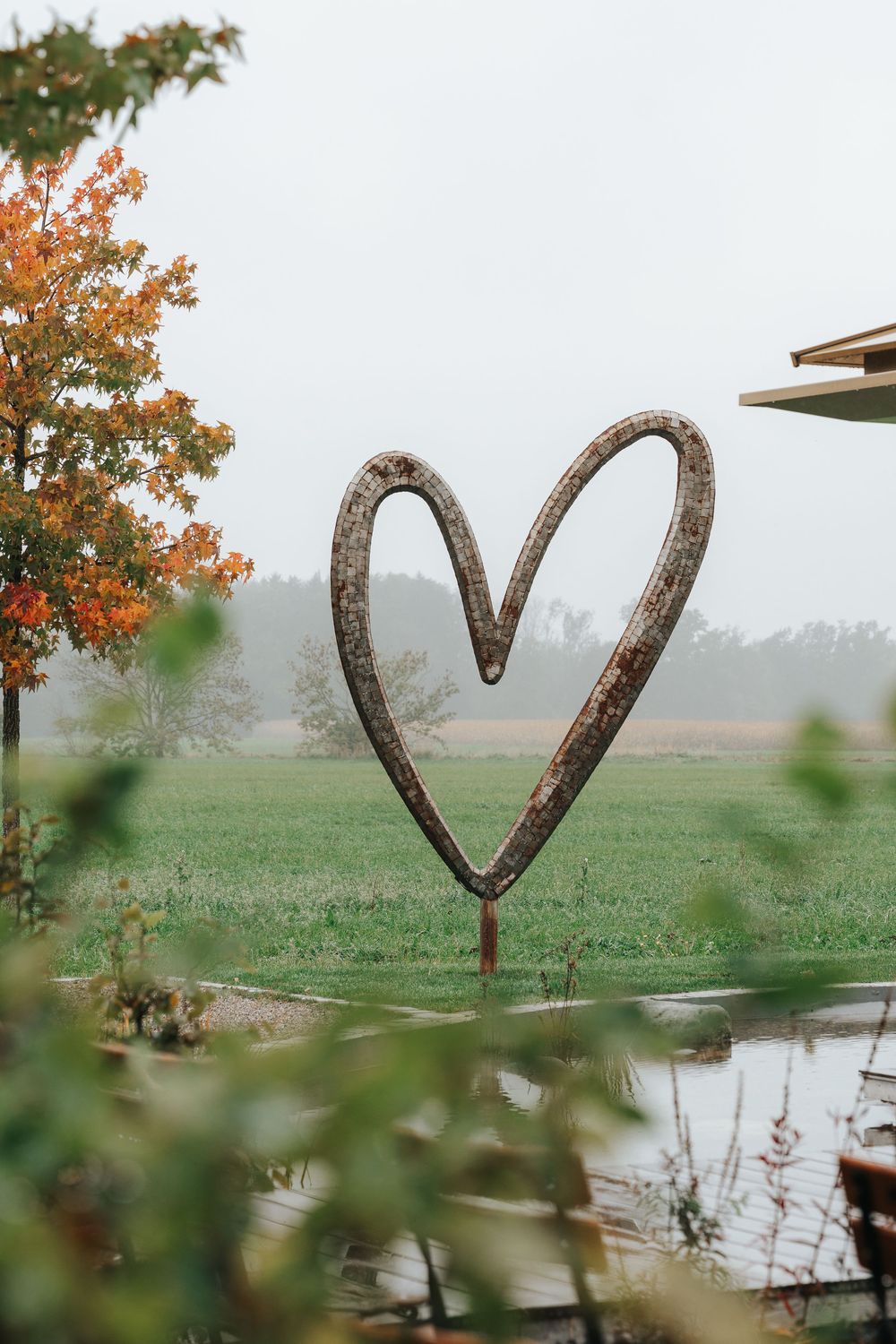 A rustic heart-shaped metal decoration hangs in front of a misty autumn landscape with orange trees and a pond.