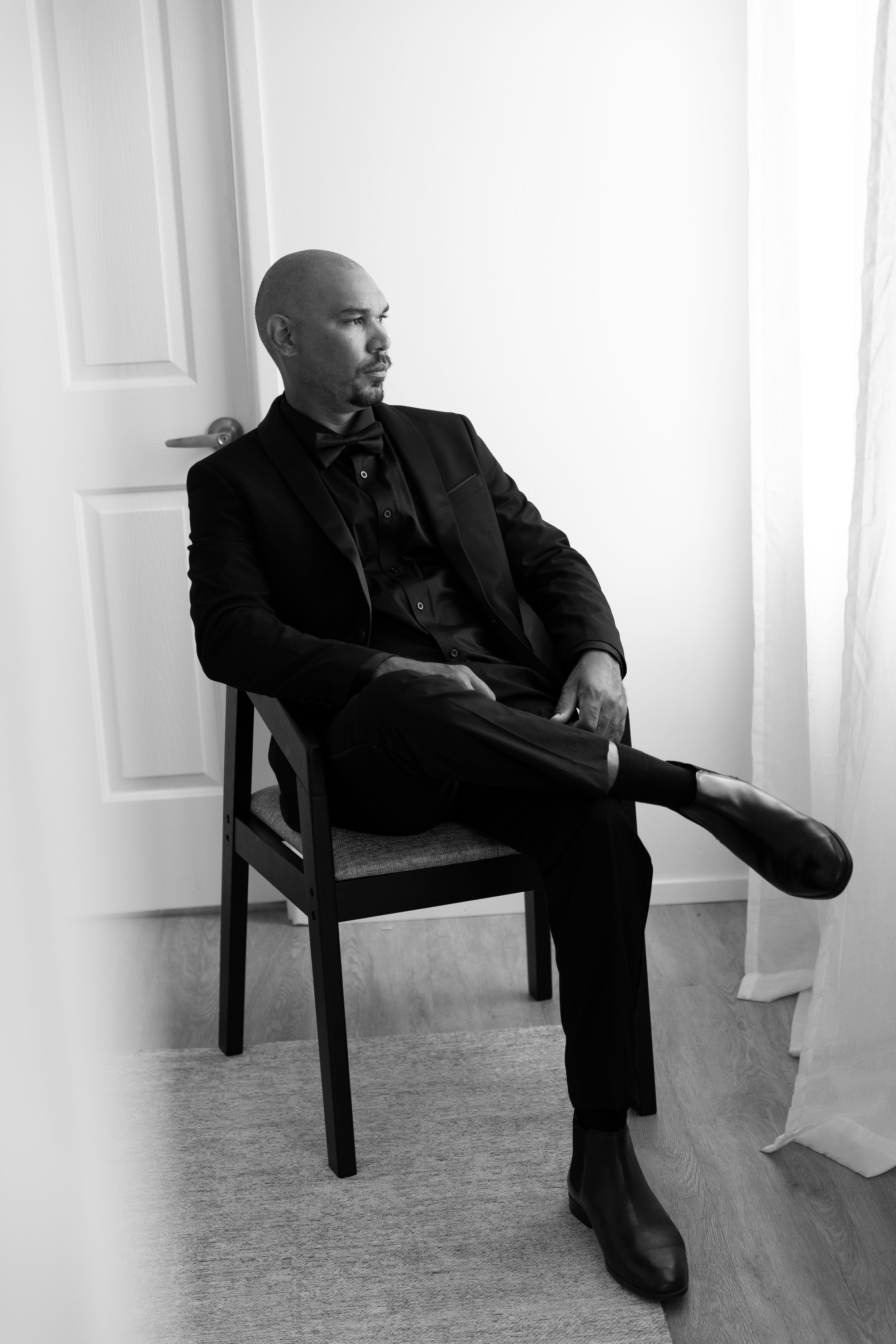 Person in black suit sitting casually on a wooden chair against white wall in artistic black and white photo.