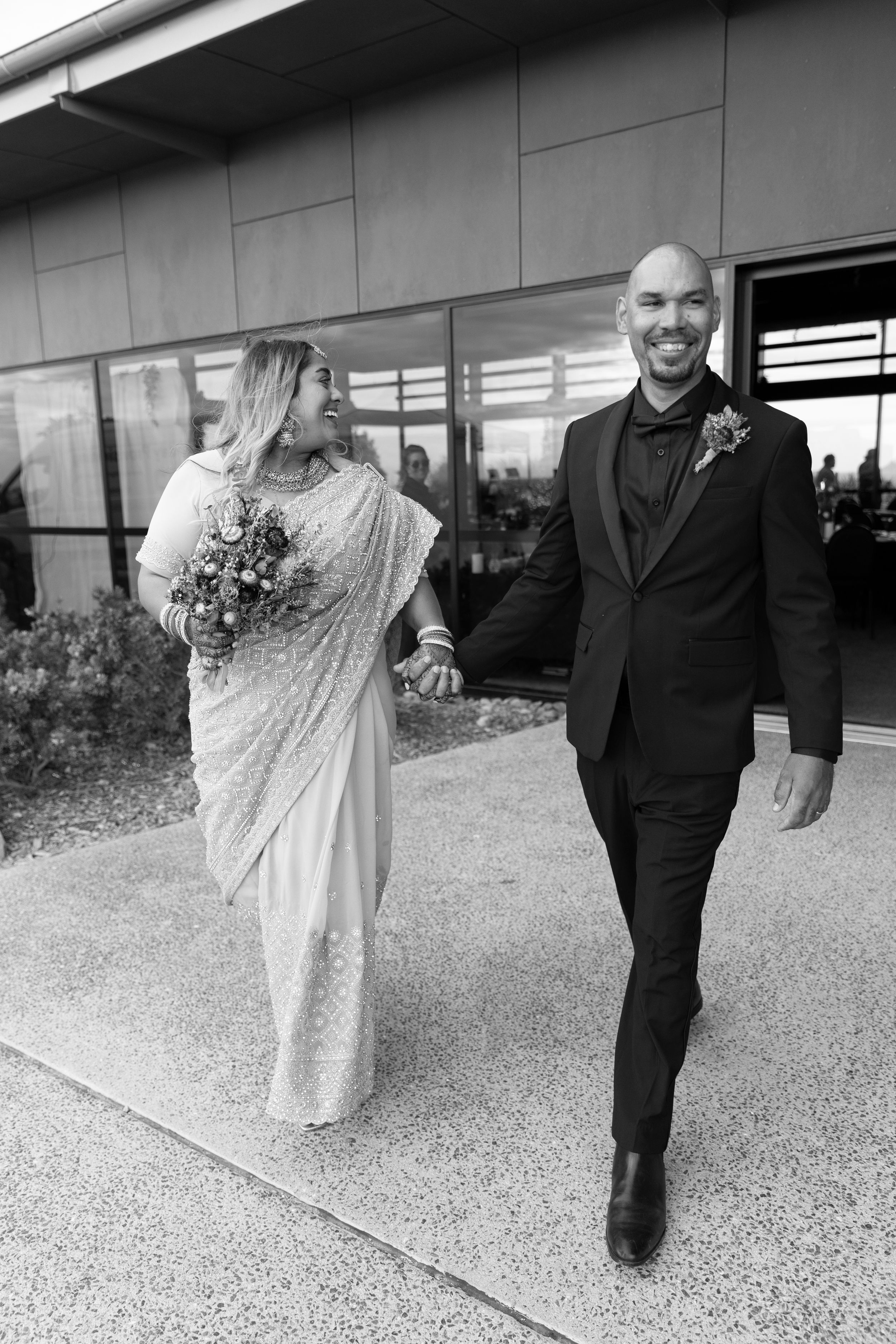 A bride in a white sequined dress and groom in black tuxedo walk together outdoors in black and white photography.
