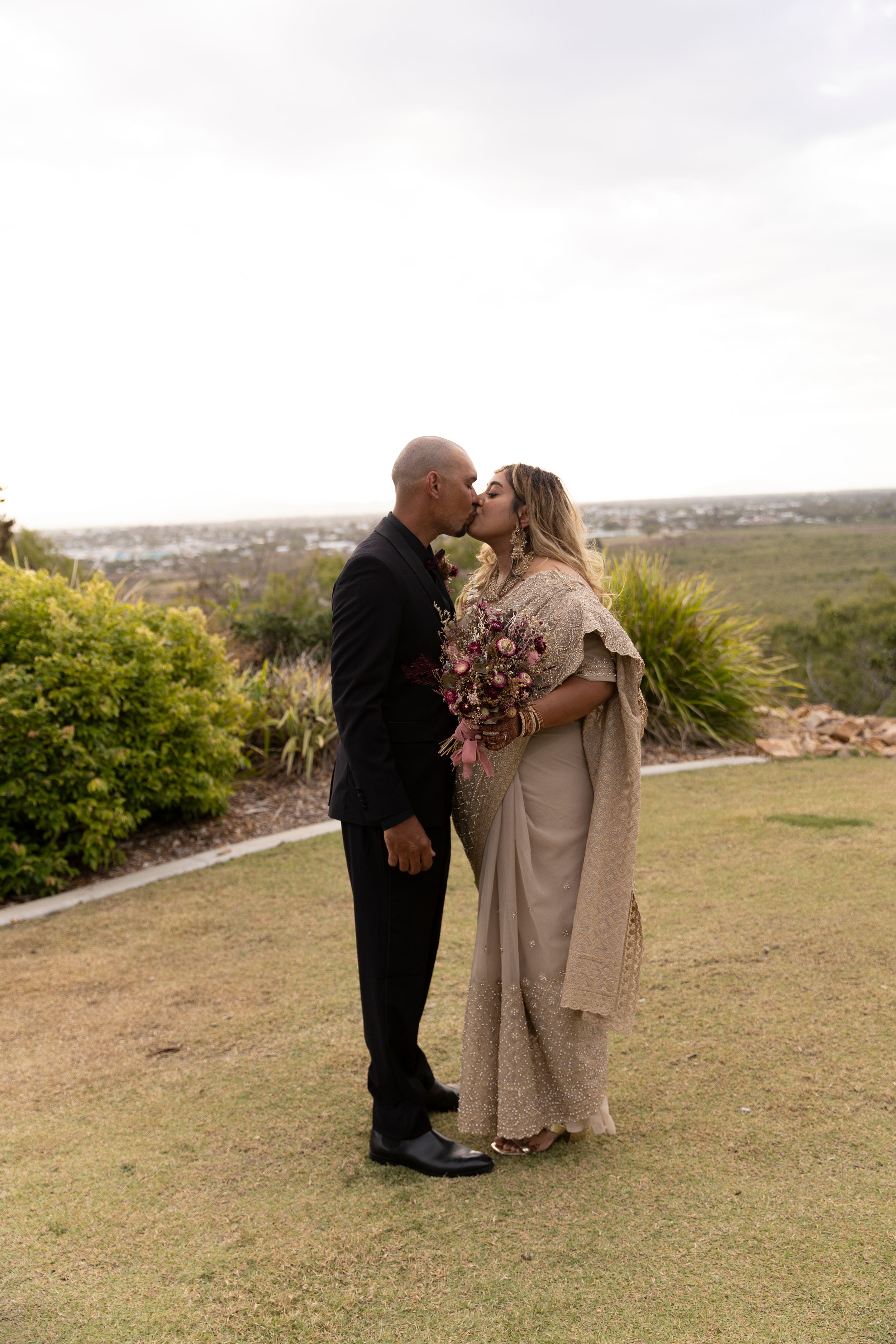 Two people in formal attire share an intimate moment on a grassy hillside overlooking a scenic landscape.