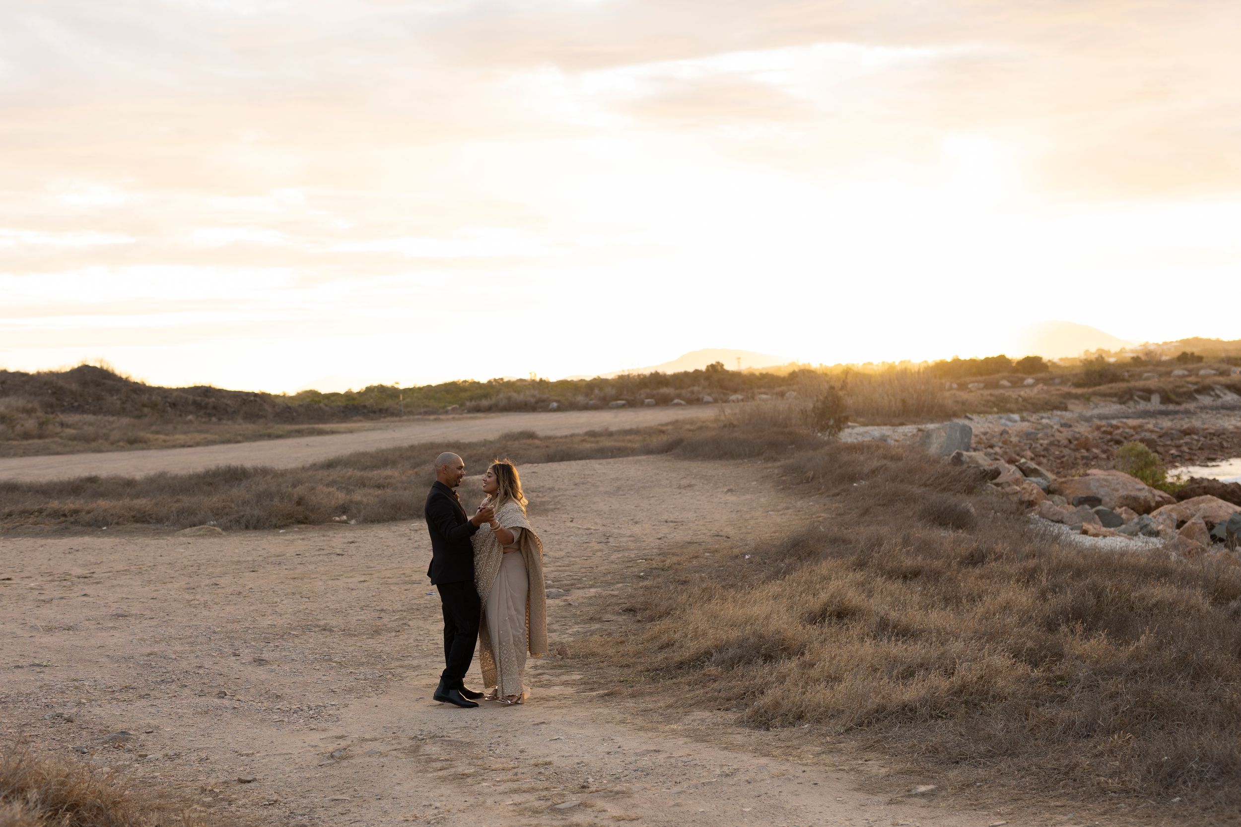 Two silhouettes embrace on a desert path at sunset with warm golden light illuminating the barren landscape.