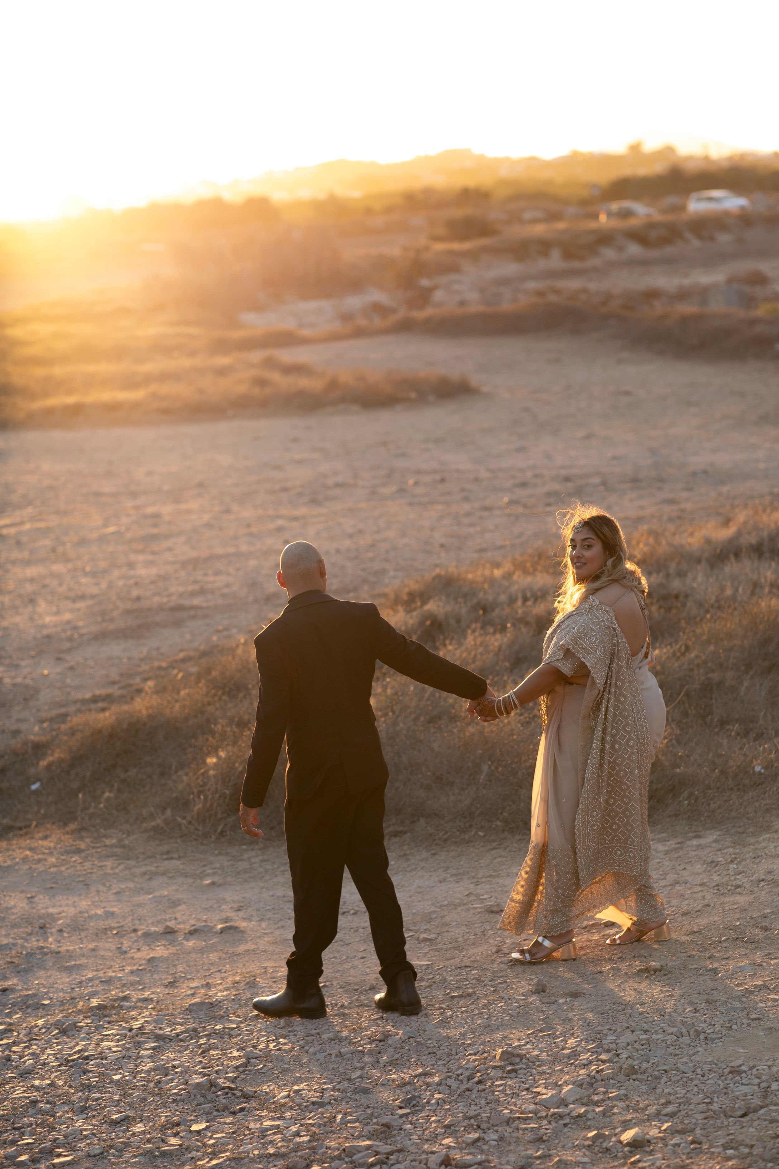 Two people hold hands and walk together on a rocky desert landscape during golden hour sunset.