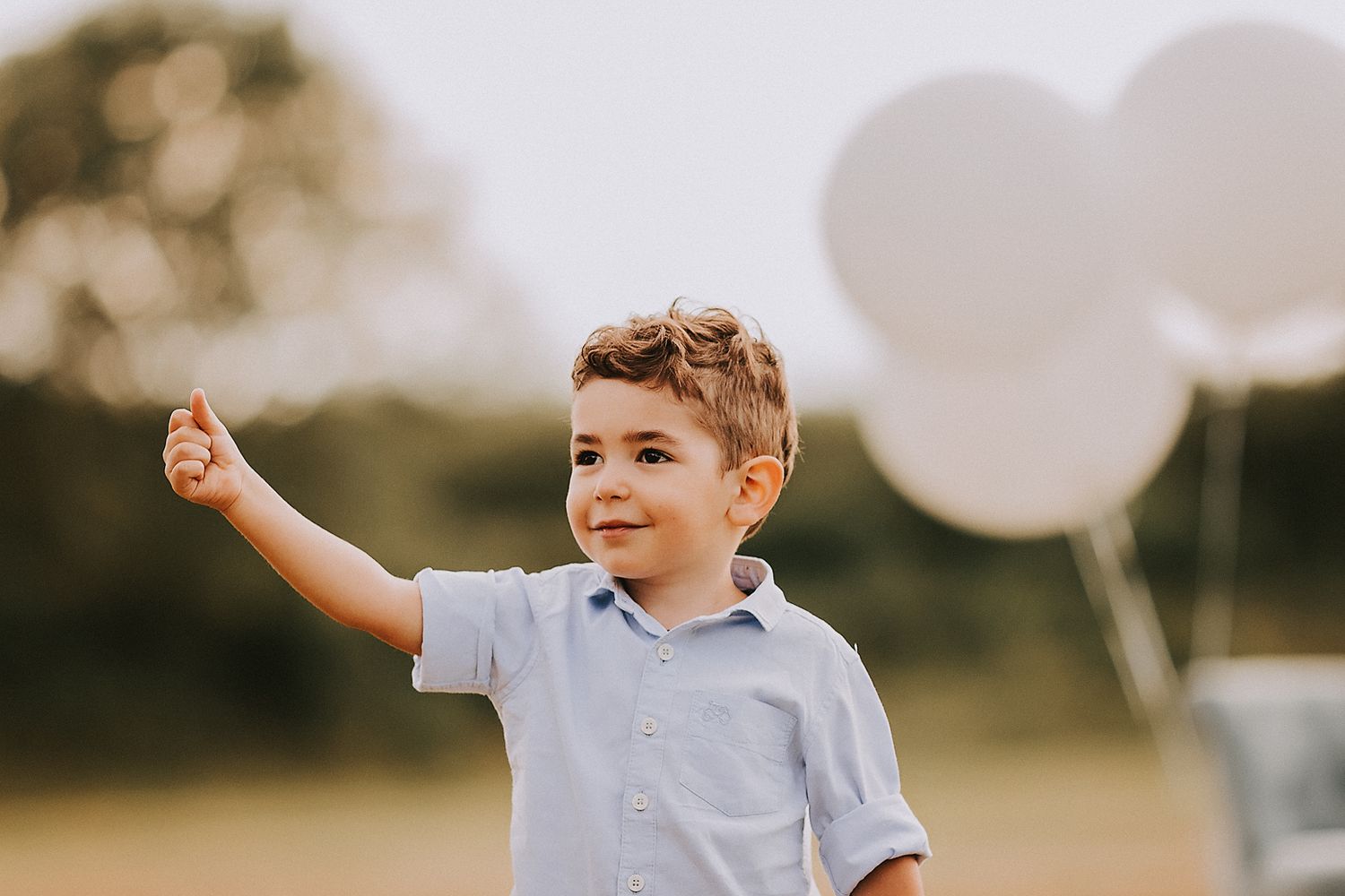Young child in light blue polo shirt giving thumbs up outdoors with white balloons in soft focus background.