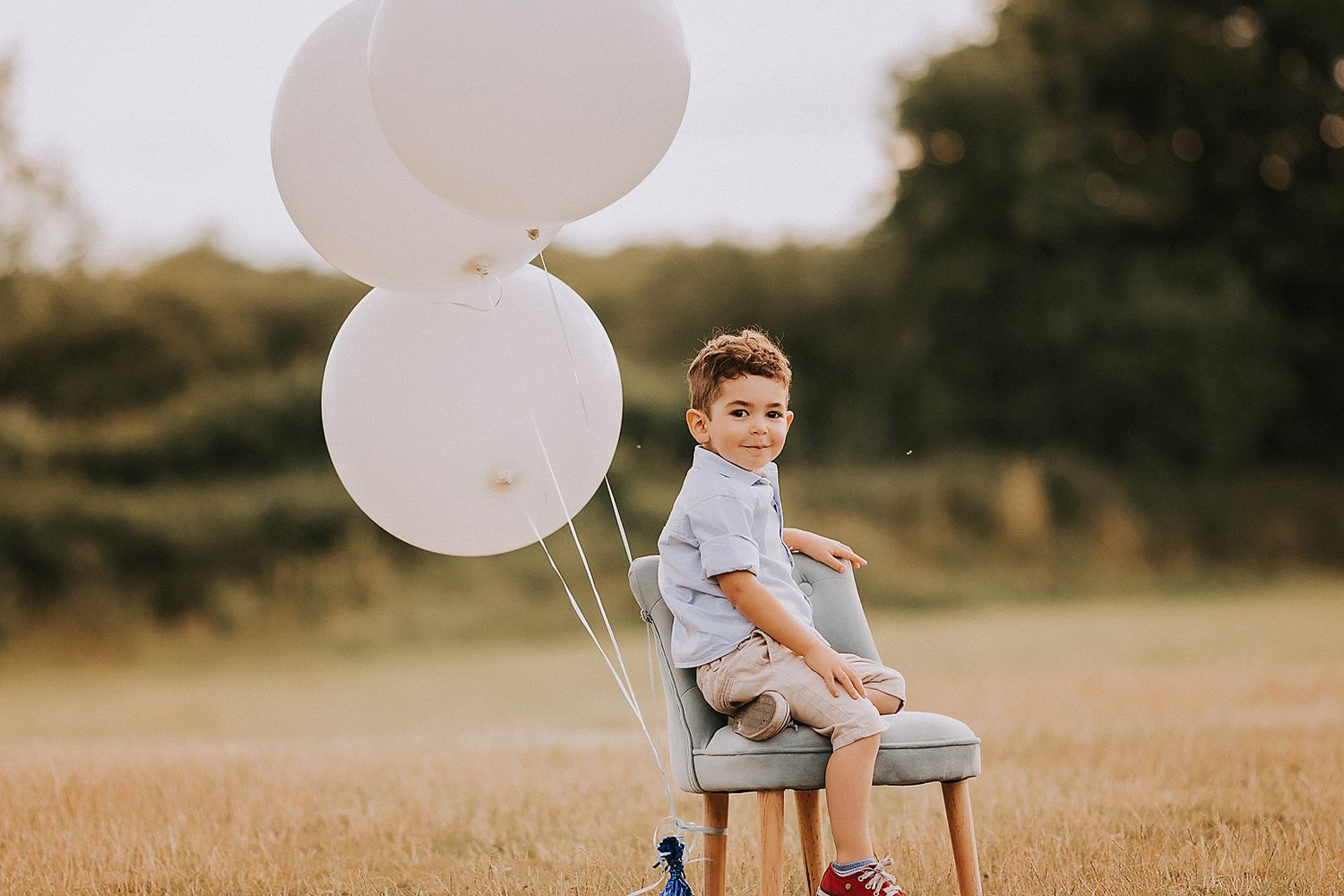 A child sits on a wooden chair in a golden field with white balloons at sunset.