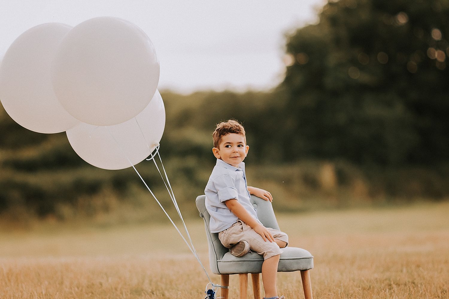 A child sits on a vintage chair in a golden field at sunset holding white balloons.