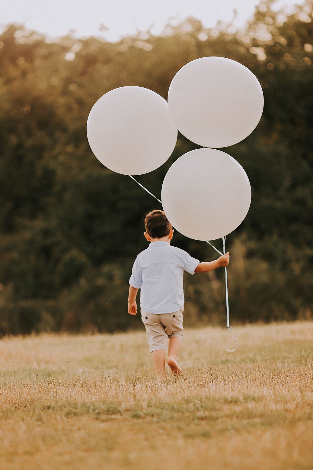 A child in a white shirt runs through a golden field at sunset holding three white balloons.
