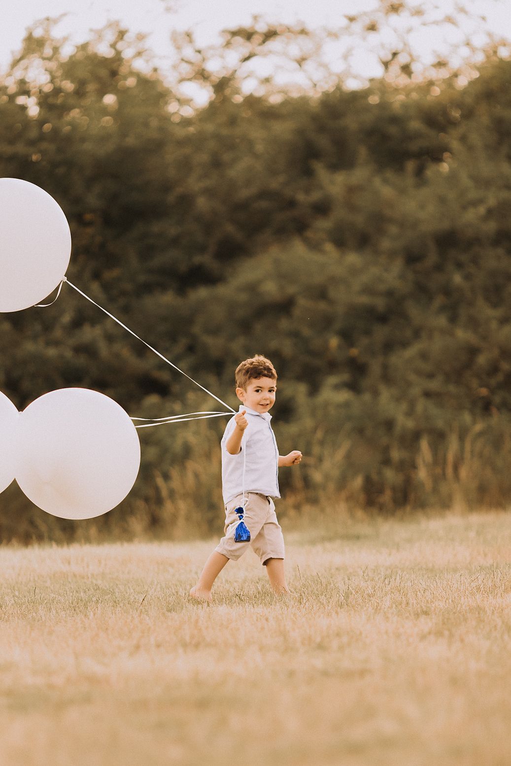Child plays with white balloons in a golden field during sunset creating a dreamy outdoor scene.