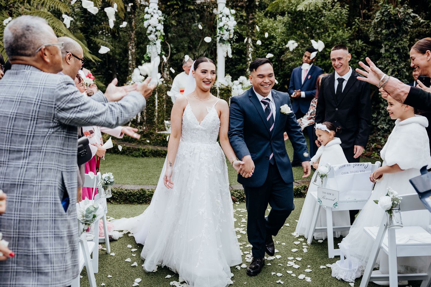Wedding guests throw confetti as a couple walks down the aisle under natural outdoor lighting.