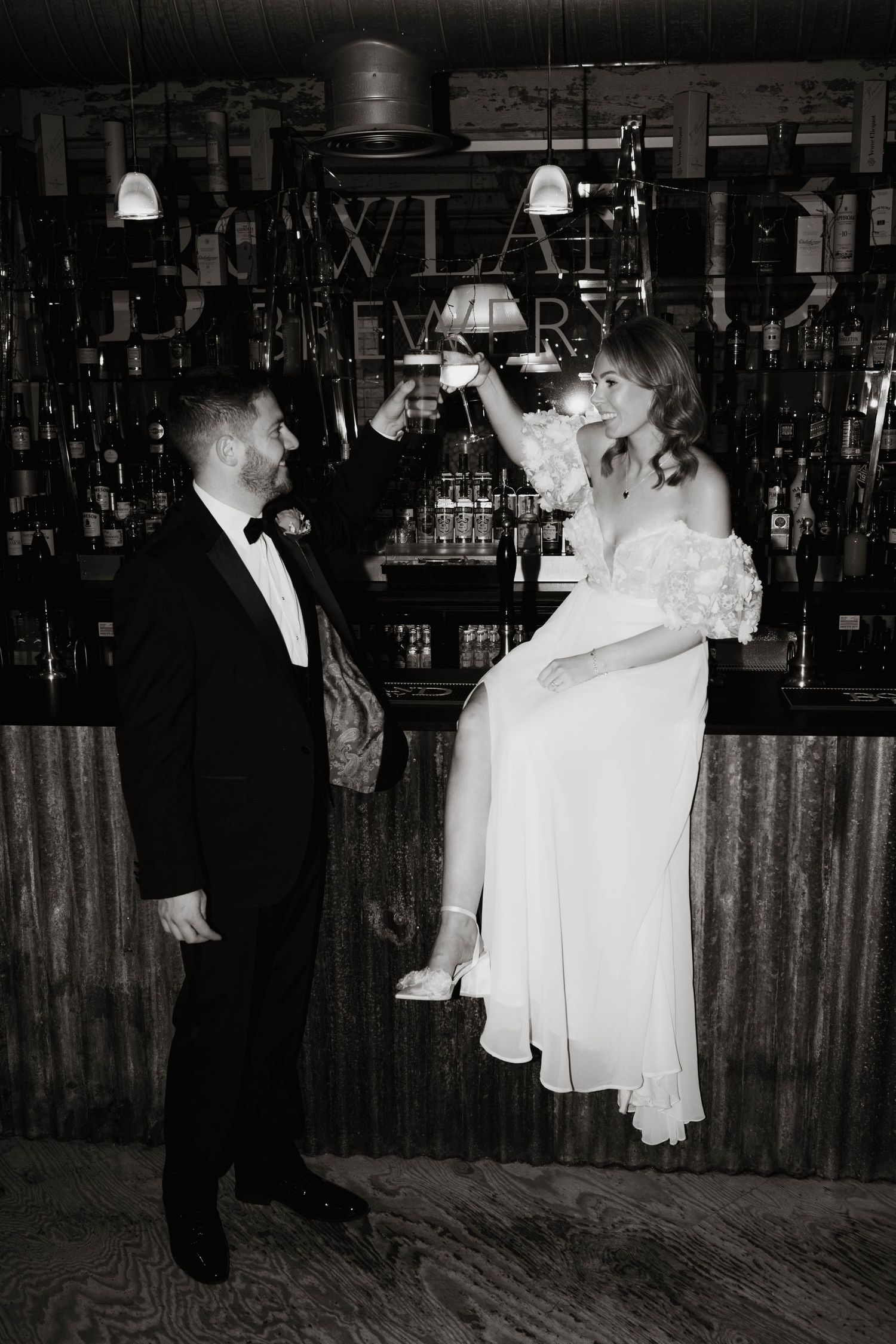 Editorial black and white wedding photo of bride sitting on bar, raising a toast with direct flash by Manchester wedding photographer