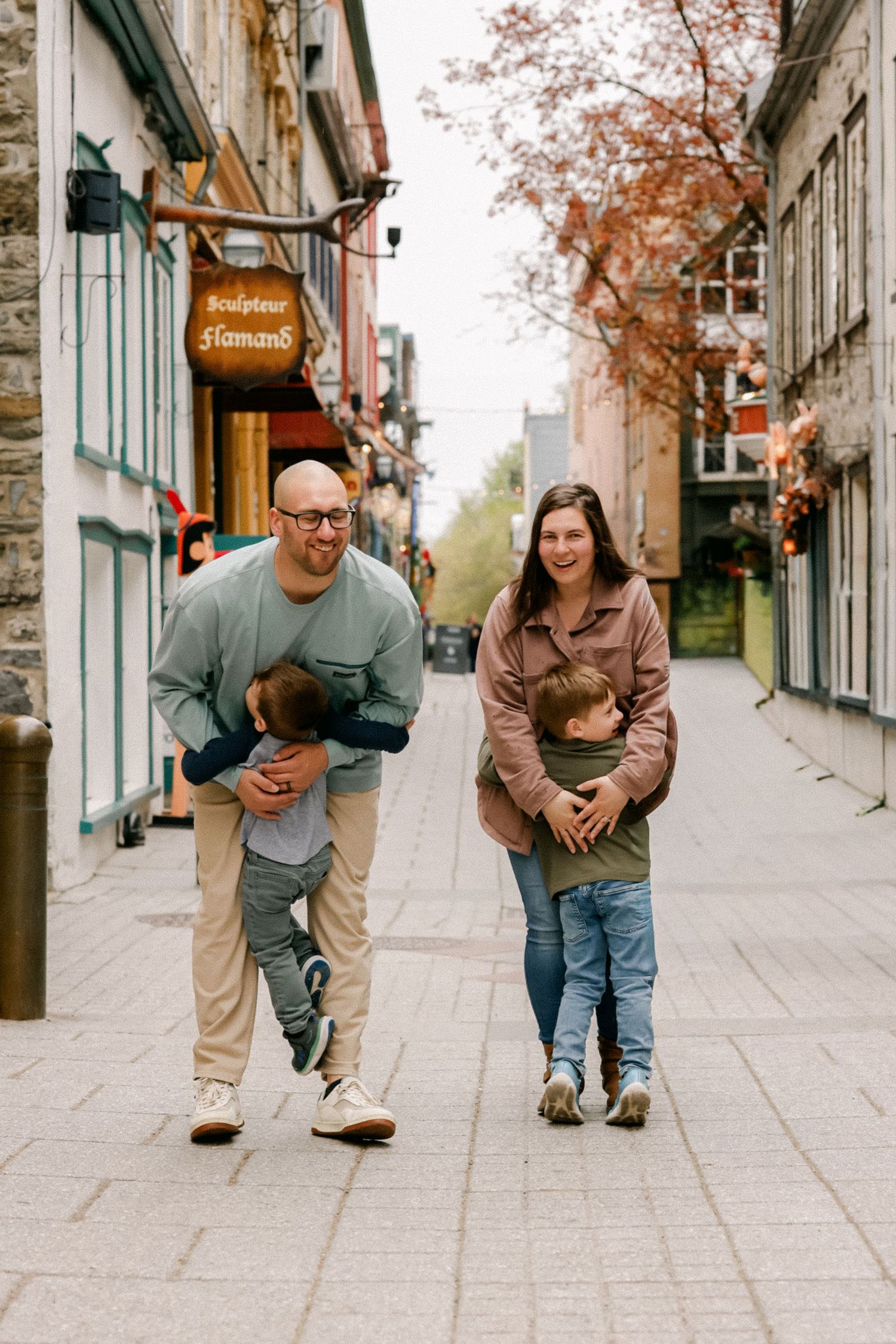Family of four walking on a cobblestone street in Old Quebec City, parents playfully holding young children outdoors.
