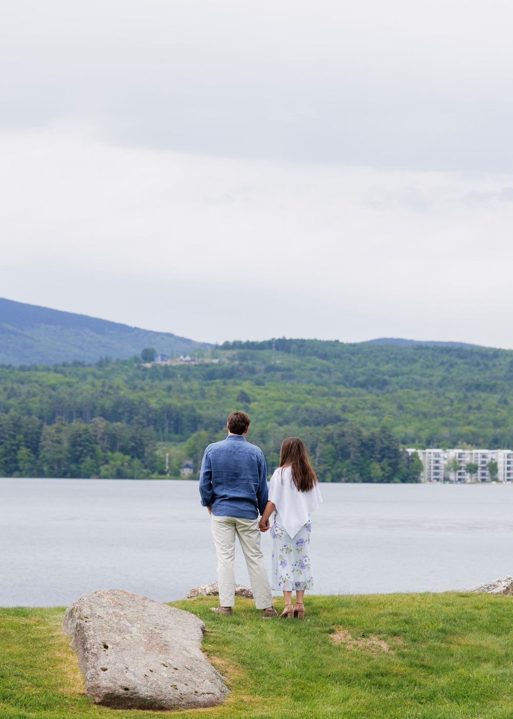 Couple holding hands standing on grassy lakeside with mountain views and cloudy sky.