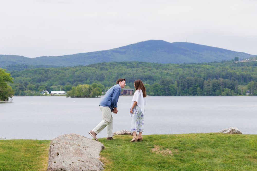 Couple holds hands near rocky lakeside with mountain and green forest backdrop on cloudy day.