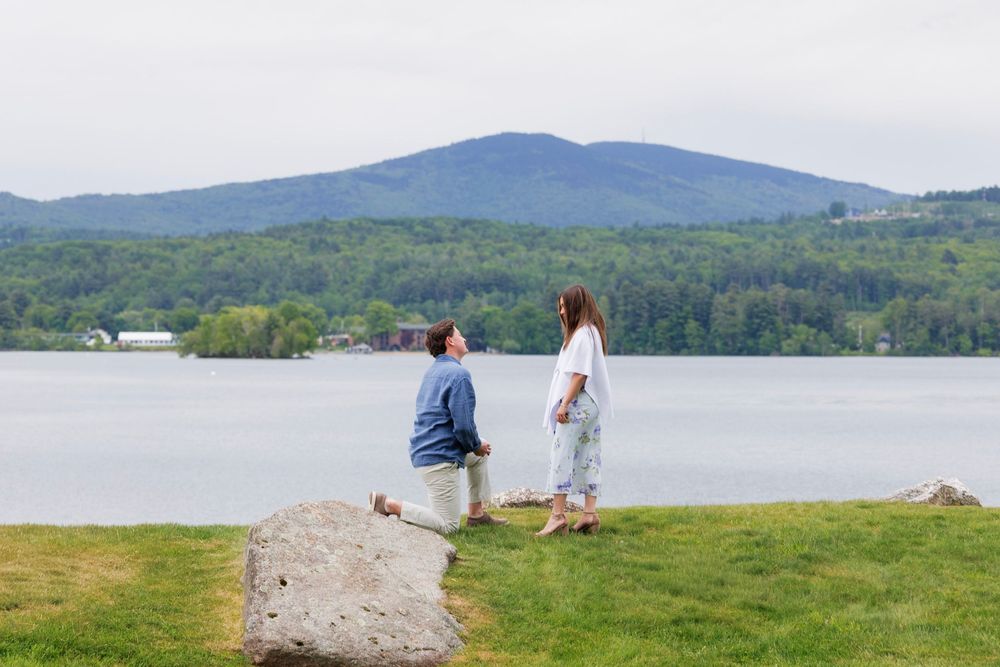 Couple standing by a serene lake with mountain views and lush green trees in the background.