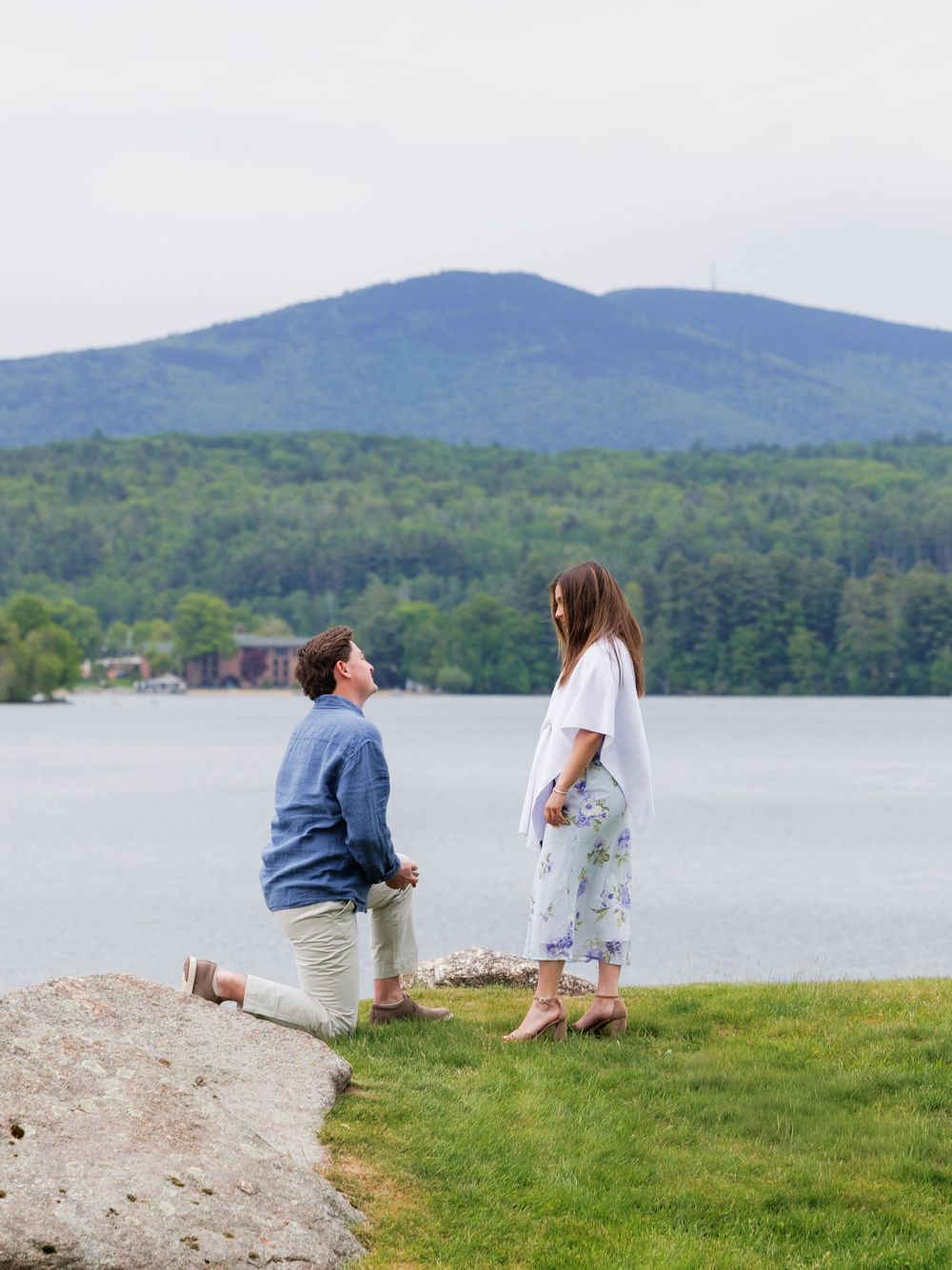 Person proposing on one knee by a lakeside with mountains in the background on a sunny day.