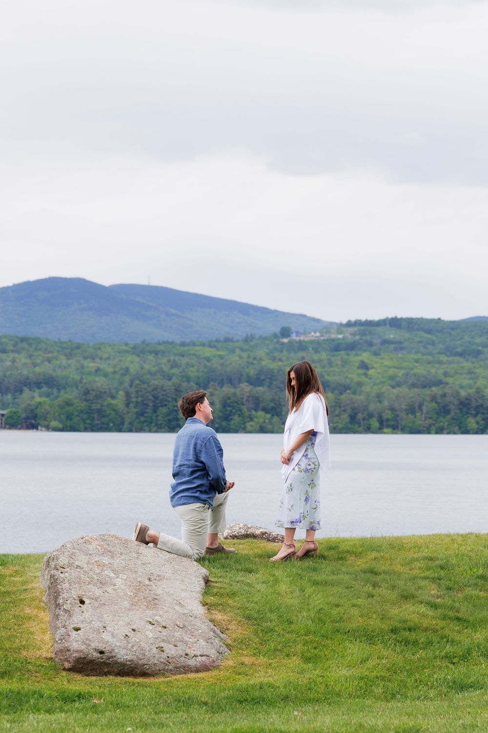 Man kneeling on one knee proposing to woman by a serene lake with mountain backdrop on a cloudy day.