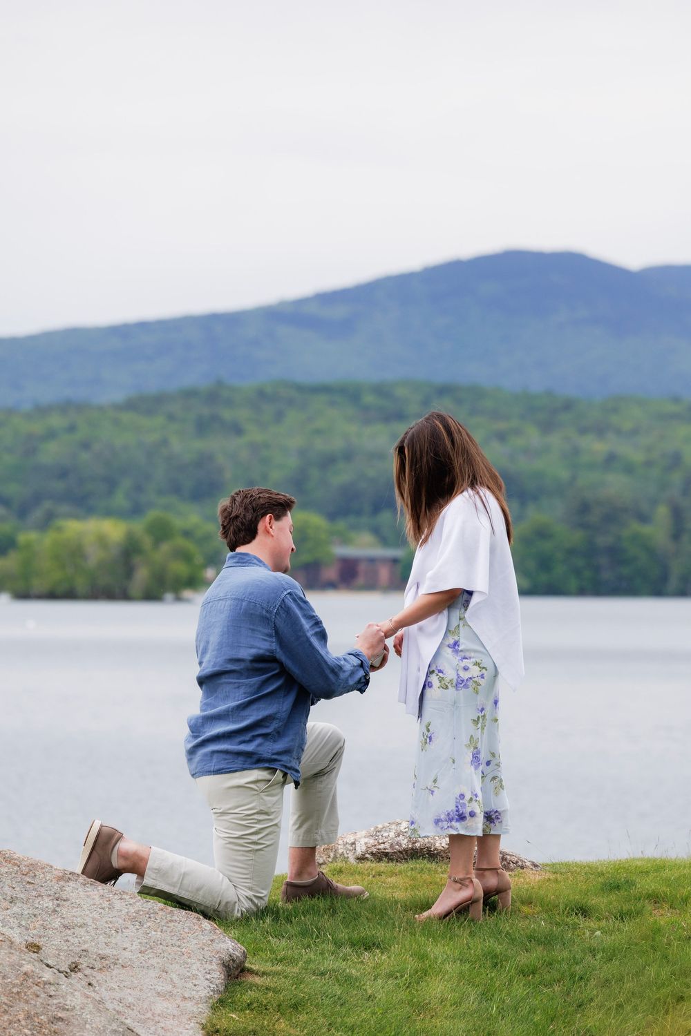 Man kneeling on one knee proposing to woman by a scenic lake with mountain backdrop.