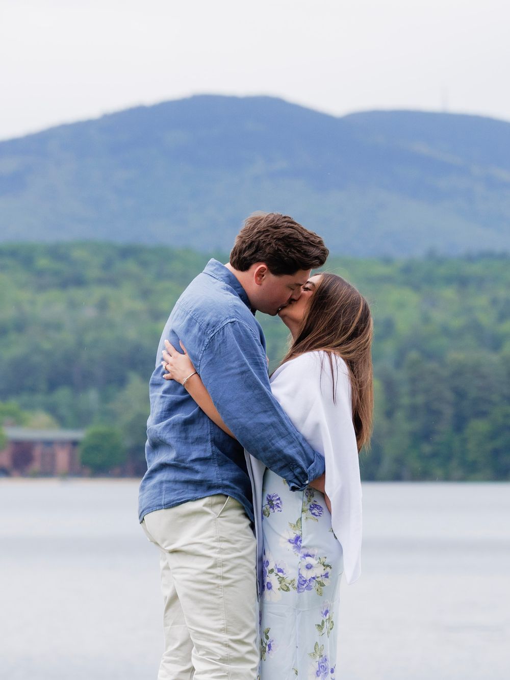 Couple sharing a romantic kiss by a lake with a mountain and green trees in the background.