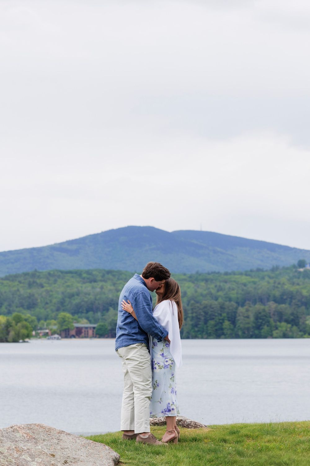 Couple sharing a romantic kiss while embracing by a peaceful lake with lush mountain scenery behind them.