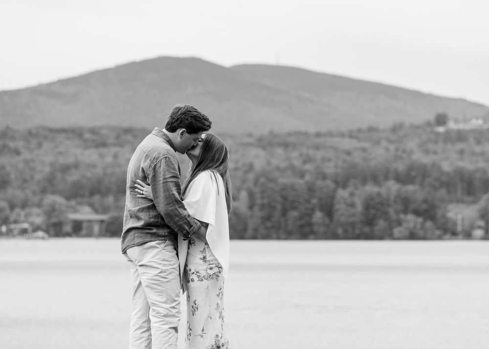 Black and white photo of a couple kissing near a lake with a mountain backdrop.