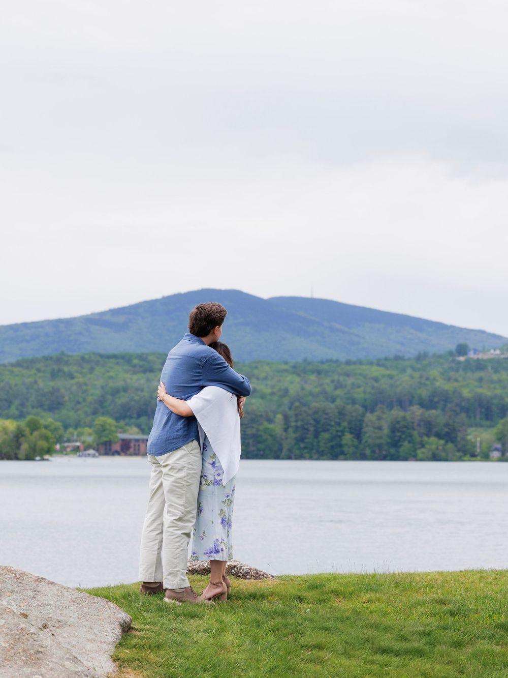 Couple embracing while gazing at a serene lake with mountain backdrop on a cloudy day.