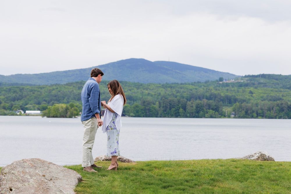 Couple stands facing each other holding hands by a lake with a mountain and forest behind them.