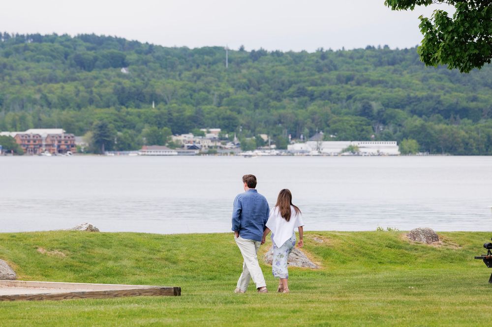 Couple walks hand in hand on green lawn overlooking a serene lake with tree-covered hills.