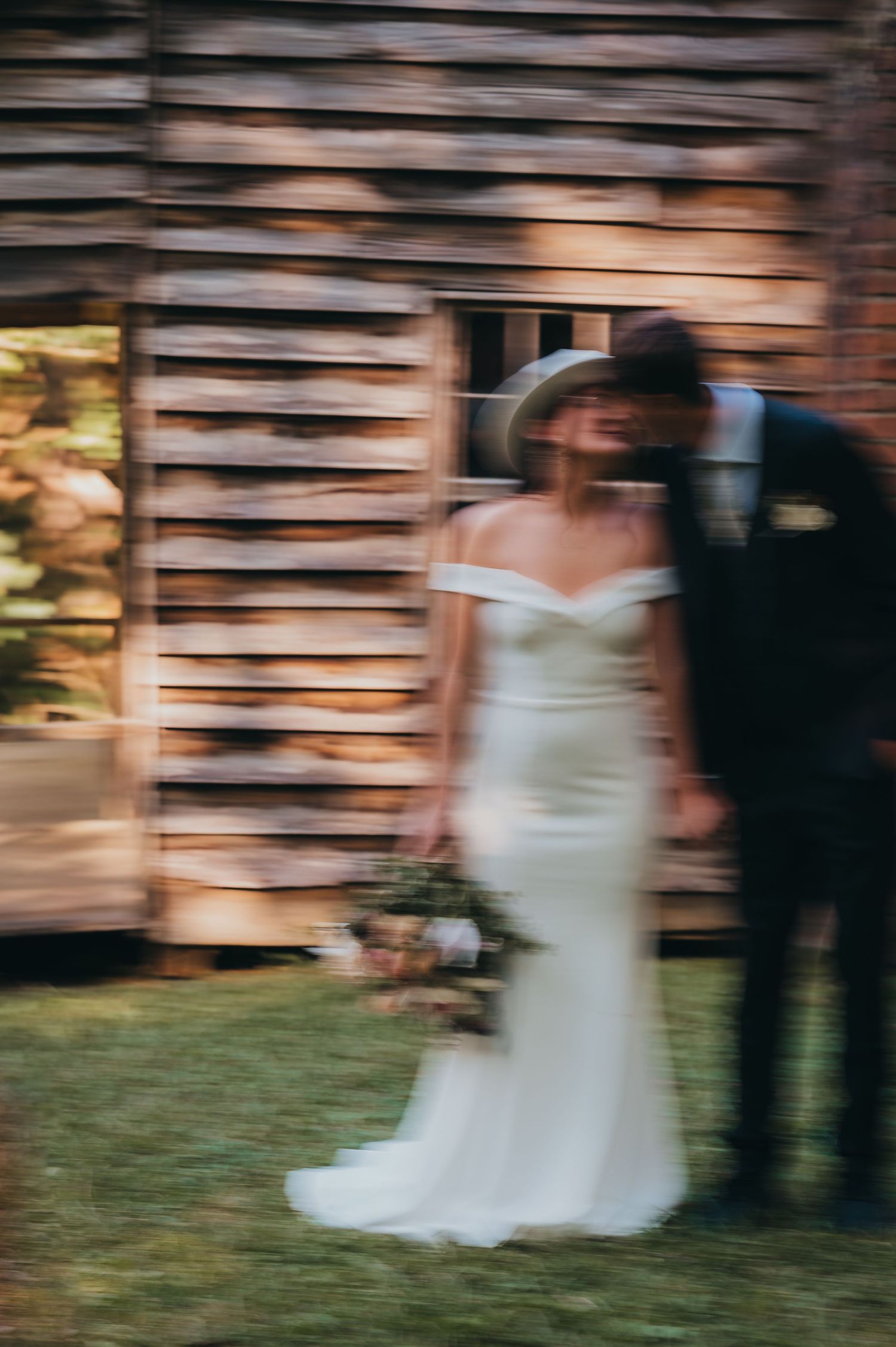 Blurry artistic wedding photo of bride in white gown and groom in suit standing before rustic wooden barn.