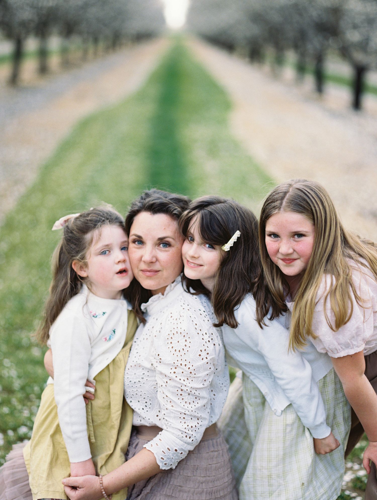A smiling mother poses with her three daughters along a tree-lined orchard path.